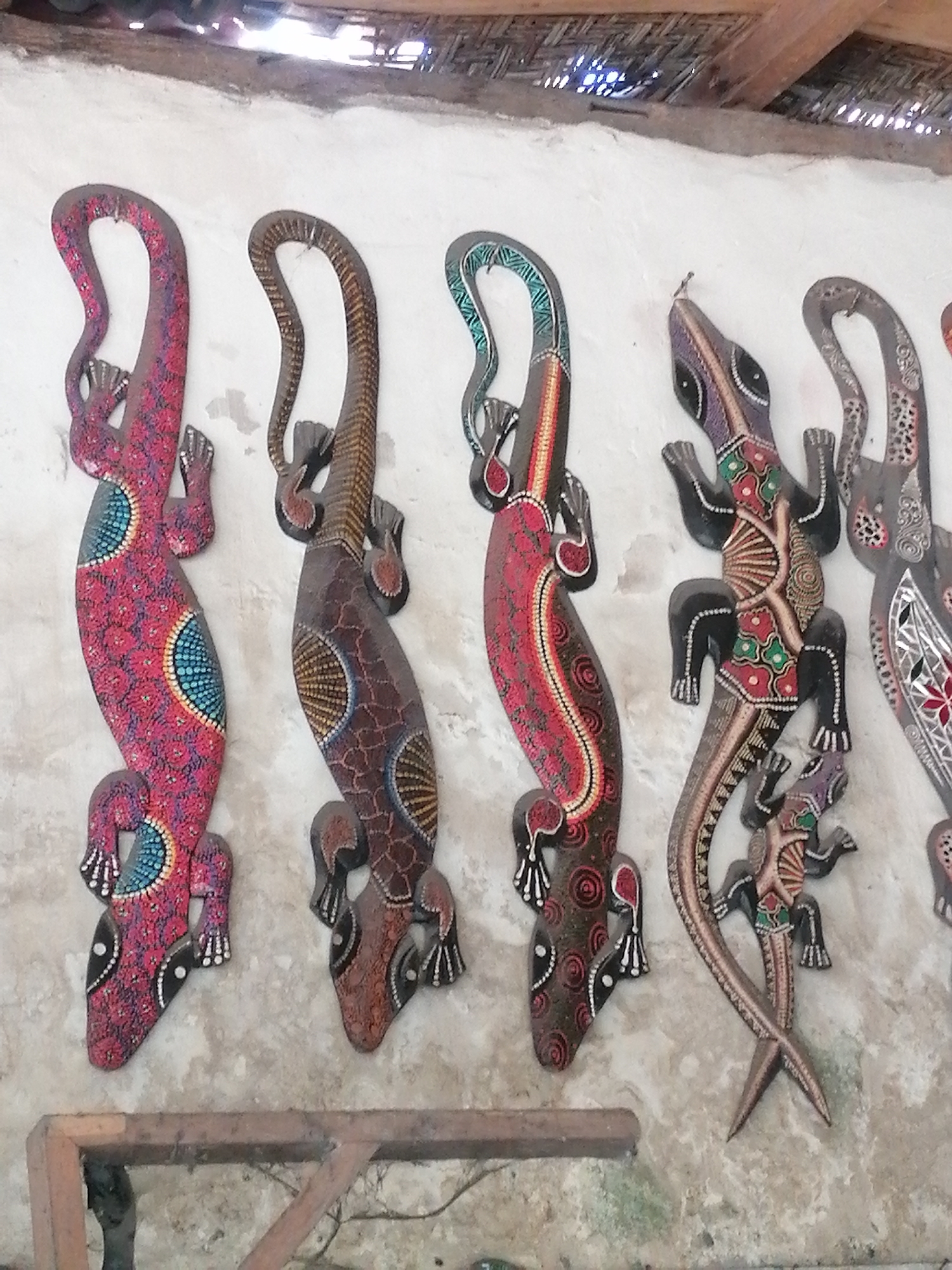 The image displays a wall adorned with five intricately carved wooden lizards, a common decorative item found in artisan shops in Ubud, Indonesia. The lizards are positioned vertically, their tails curving upwards, creating a visually dynamic arrangement. Each lizard is uniquely painted with vibrant patterns, featuring a combination of red, yellow, blue, and green hues, with intricate dotwork and geometric designs adorning their bodies. The first lizard from the left is predominantly red with blue circular patterns. The second is brown with yellow and brown markings. The third lizard is painted with green and blue scales along its back. The fourth, and largest, exhibits a more complex pattern of red, green, and black. The fifth lizard, partially visible on the right, shares a similar style with the fourth. The wall itself appears to be made of rough, textured plaster, with a wooden beam or shelf visible at the bottom of the frame. Above the lizards, a thatched roof structure is visible, suggesting an open-air setting or a building with traditional Indonesian architecture. The lighting indicates daytime. The overall scene is characteristic of a craft market or souvenir shop in Ubud, a region renowned for its traditional arts and crafts.