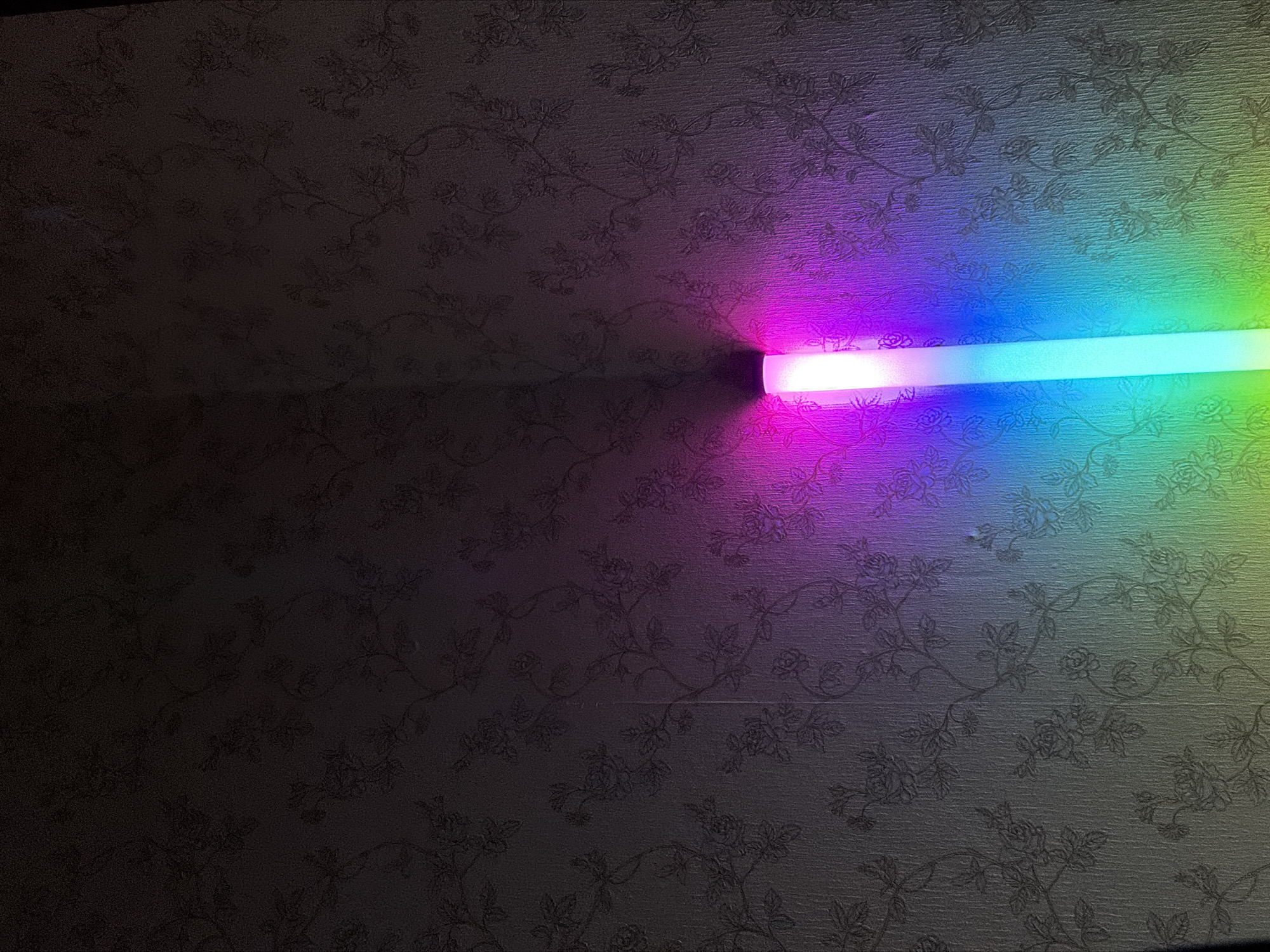 The image shows a close-up of a wall with floral wallpaper. A vertical, illuminated light fixture is positioned against the wall, casting a gradient of colors from pink and purple at the top to blue and green towards the bottom. The light is casting a shadow onto the wall. There are no people or animals visible in the image. The scene appears to be indoors.