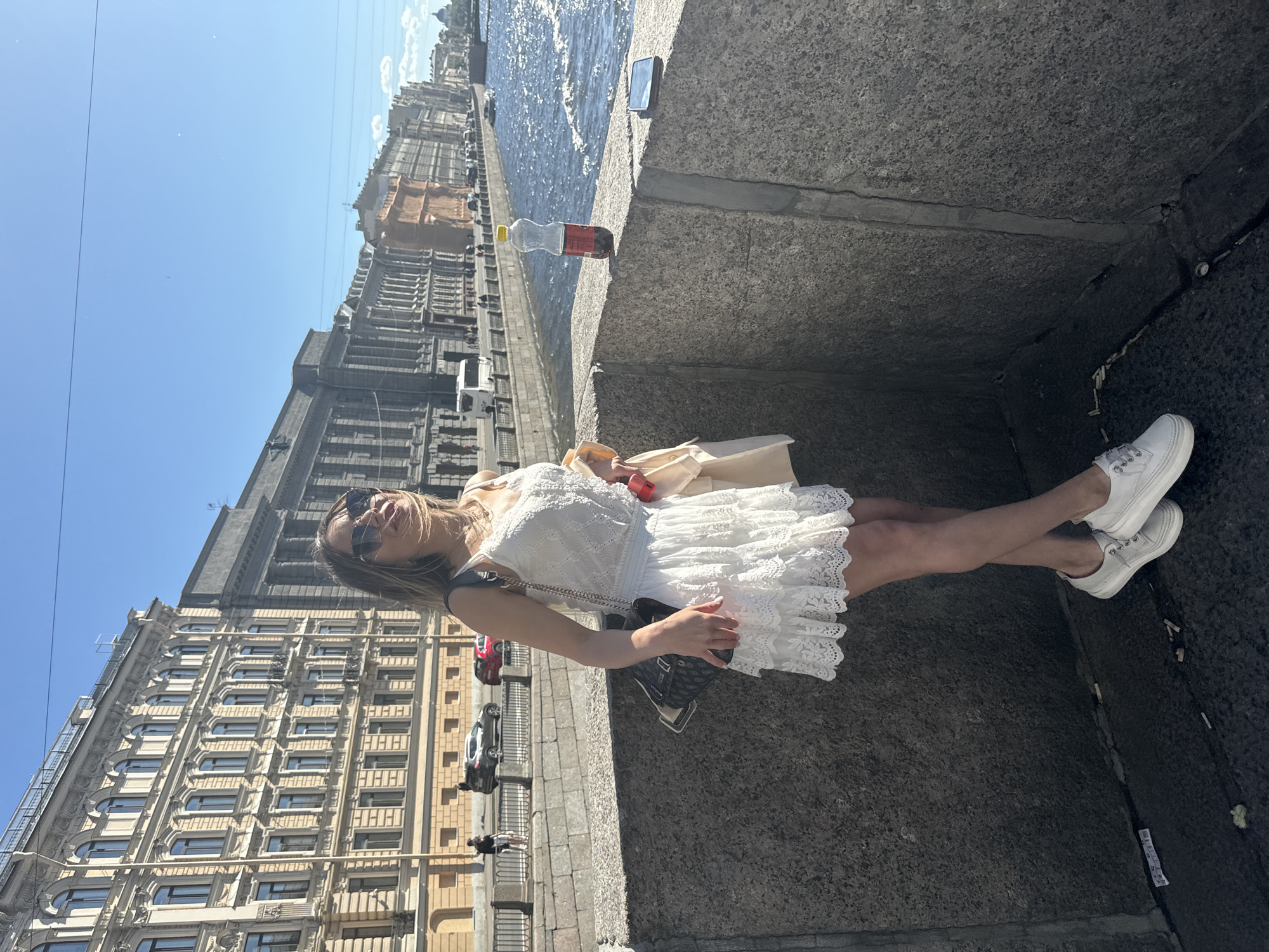 The image shows a woman in a white lace dress and white sneakers standing on a stone embankment by a body of water. She is wearing sunglasses and has her hair down. She is holding a beige bag and has a dark purse on her shoulder. A bottle of soda and a smartphone are on the embankment next to her. In the background, there are buildings with many windows and cars on a street. The sky is blue and it appears to be a sunny day. The scene suggests a casual outing or a tourist enjoying the city. The buildings and the architectural style are consistent with a European city.