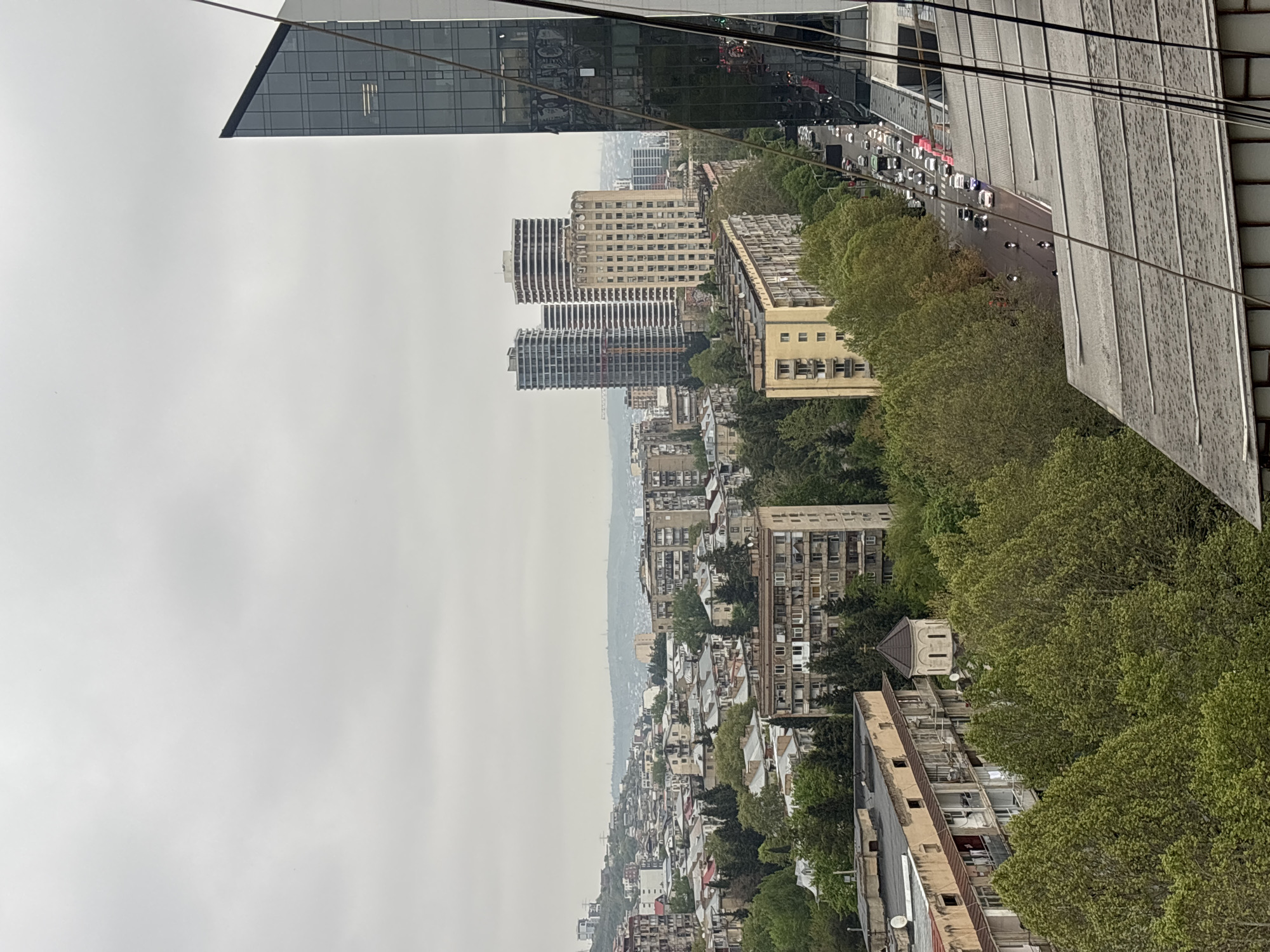 This image captures a cityscape in Tbilisi, Georgia, under an overcast sky. Tall, modern buildings are seen in the distance, alongside older, Soviet-era apartment blocks, creating a contrast in architectural styles. A tree-lined street with light traffic flows parallel to the buildings. The photo seems to have been taken from an elevated position within a modern glass building, with parts of the structure visible in the frame. The overall environment suggests an urban setting with a blend of older and newer construction.