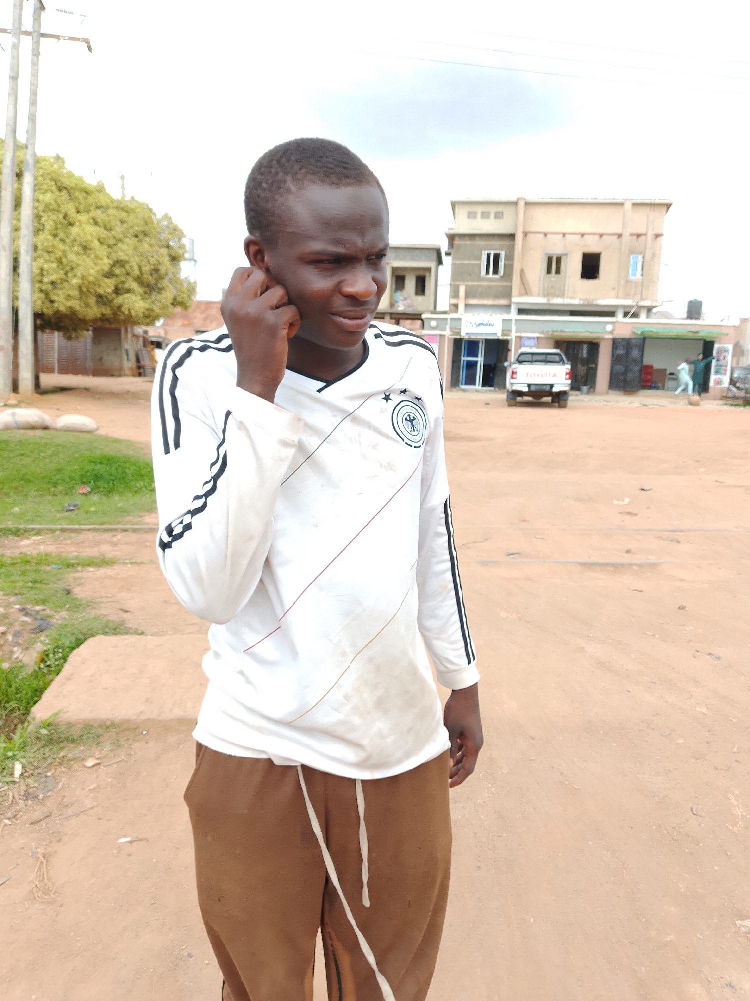 A young man is standing outside and looking to his left with his hand to his ear. He is wearing a white long-sleeved shirt with black stripes on the sleeves and brown pants. In the background, there are buildings and a white pickup truck. The ground is dirt and there is some grass and a tree to the left. The sky is cloudy.