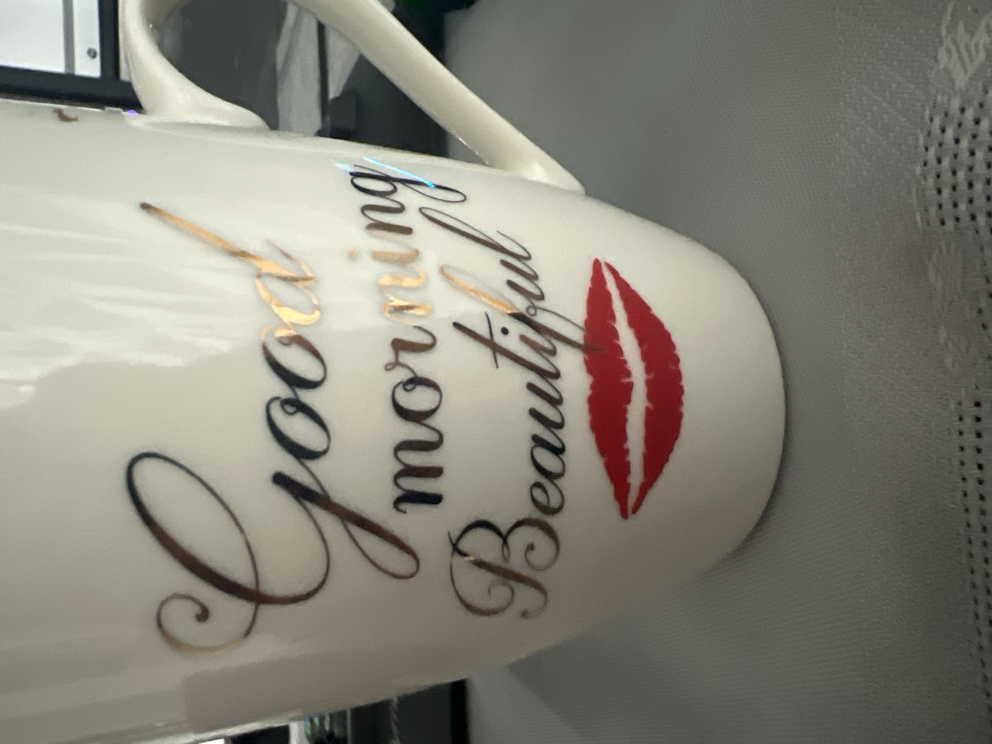 A close-up, slightly angled view showcases a white ceramic mug with the phrase "Good morning Beautiful" written in a decorative cursive font. The text is primarily in black with golden accents on the edges of the letters, except for the word "Beautiful," which is completed with a red lipstick mark to represent a kiss. The mug has a white handle that curves smoothly. The background consists of indistinct gray surfaces, possibly a table or desk, with a fine mesh-like texture partially visible.