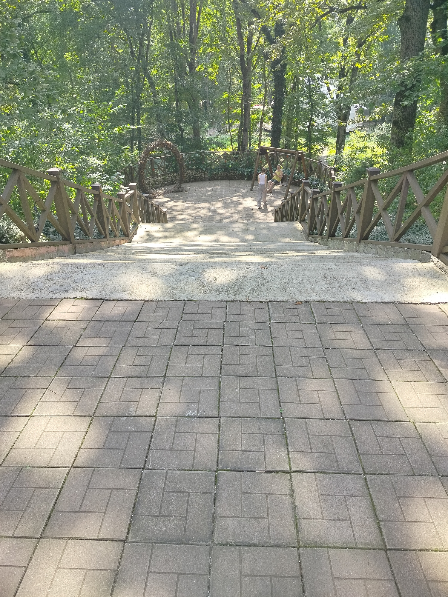 This image depicts an outdoor scene in the city of Krasnyi Most, Russia. The foreground is dominated by a paved pathway made of interlocking gray square tiles, with a herringbone pattern. Sunlight casts distinct shadows across the tiles.
A set of concrete stairs descends from a landing at the top of the tiled pathway. These stairs are flanked by a wooden railing with a criss-cross design, supported by vertical posts. The railing follows the gentle slope of the path leading into a wooded area.
The landing at the top of the stairs appears to be a paved area with a different, lighter-colored material. Two individuals are visible on this landing. One person, wearing light-colored pants and a white top, is walking away from the camera. Another person, in a yellow top and light-colored shorts, is seated on a wooden swing set to the left of the path.
Beyond the landing, the path continues through a park-like setting with mature trees and lush green foliage. A large, circular decorative arch made of woven branches stands to the left of the path, further into the wooded area. The overall environment suggests a public park or recreational space on a sunny day.