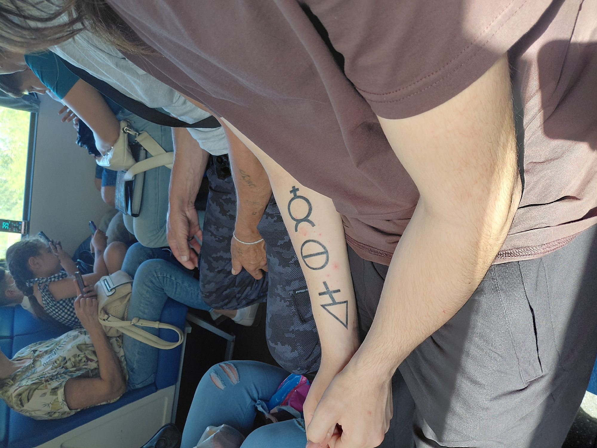 The image shows an indoor scene, likely a mode of public transportation, with several individuals seated. The primary focus is on the tattooed arm of a person in the foreground, displaying several symbols. To the left of this arm, another person's tattooed arm is visible. In the background, other passengers are seated, some holding cell phones, and a window shows a blurry outdoor environment with greenery. The lighting suggests daytime. No specific location in Novosibirsk, Russia, can be identified from the image.