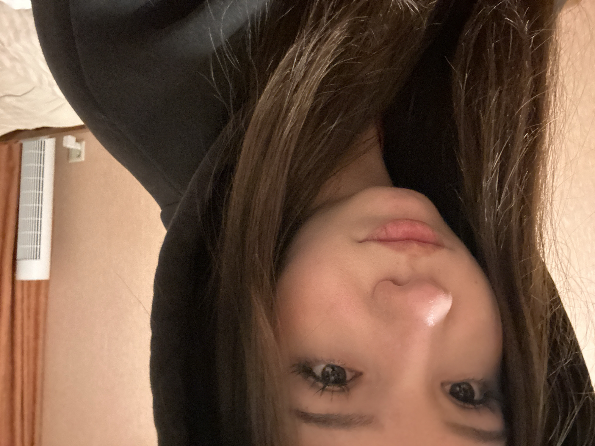 A young individual, appearing female, with long, dark brown hair and fair complexion, is depicted from the chest up, looking directly towards the viewer. She is wearing a black hooded sweatshirt with the hood drawn over her head, framing her face. Her dark eyes, enhanced with prominent eyelashes, are open. Her nose shows a highlight on the bridge, and her lips are closed in a neutral expression. The background on the left and central portion consists of a plain light peach-colored wall. To the right, an orange or burnt orange curtain is visible, along with a tall, white, vertical appliance, likely a fan or heater, with a visible grate. Below and to the left of this appliance, a white electrical outlet is mounted on the wall. A light-colored, possibly cream or white, quilted fabric, resembling a bedspread, is partially visible in the bottom right corner.