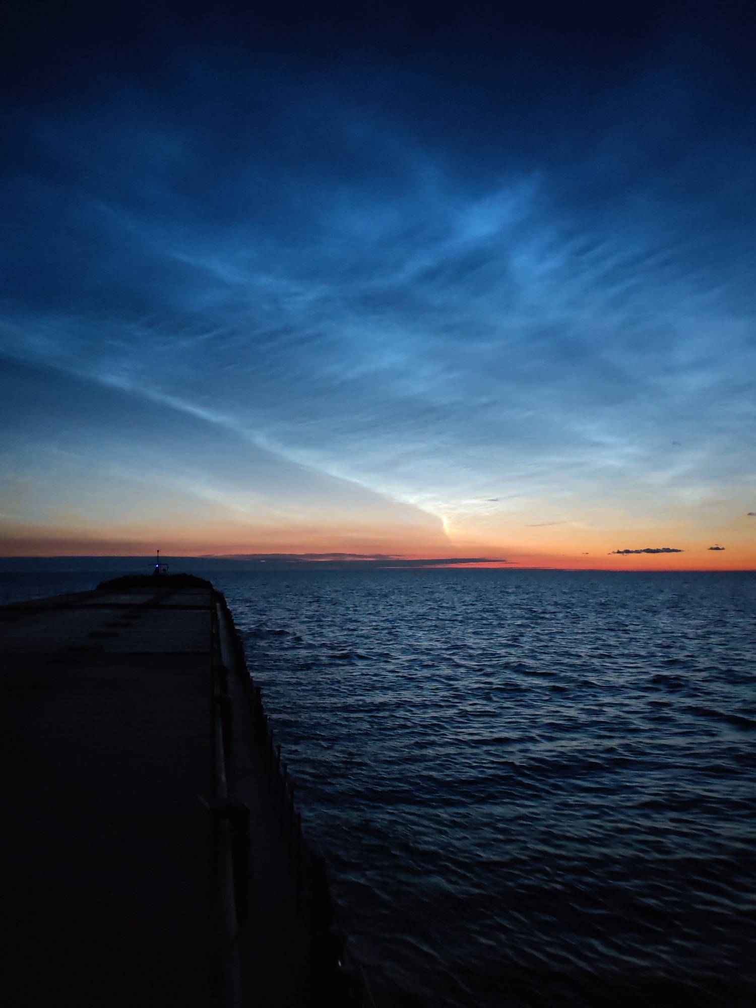 An aerial view shows a dark, stone pier extending into a dark blue, rippling ocean under a deep blue sky. The sky transitions to lighter blues and oranges near the horizon, with wispy, light-colored clouds visible. There is a small, lit structure at the end of the pier. The scene is a natural, outdoor environment at dusk or dawn. The location is identified as Unknown, Russia. There are no people visible in the image.