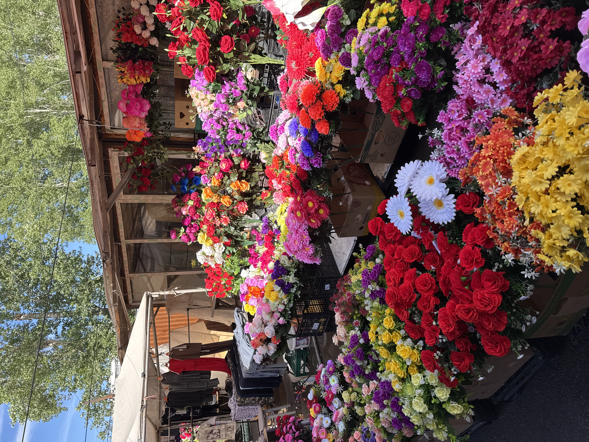 The image shows a vibrant display of artificial flowers at an outdoor market or stall in an unidentified city. The flowers are arranged in dense clusters, showcasing a wide spectrum of colors, including reds, pinks, purples, yellows, oranges, and whites. Some of the flower types include roses, gerbera daisies, and other various floral shapes. The flowers are displayed in cardboard boxes and plastic crates. The structure of the stall is visible, showing a wooden frame with a canopy and building. Hanging clothes are displayed on the side. A large tree stands near the stall. The overall impression is a colorful and bustling commercial setting.