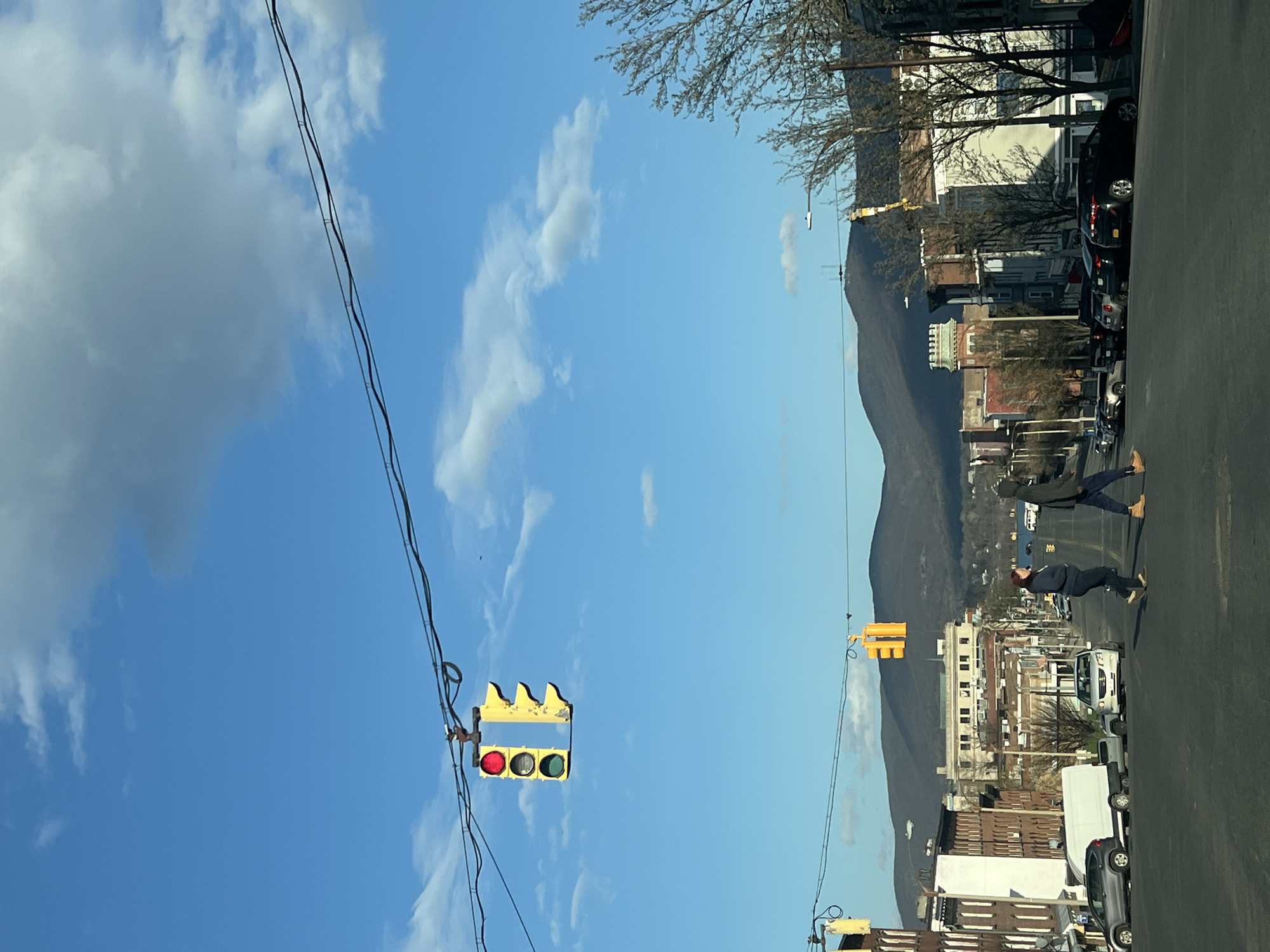 The image shows a street scene in the City of Newburgh, United States of America. A street runs through the scene with parked cars on the right side. Two people are seen crossing the street at a crosswalk. Traffic lights are present along the street, with wires stretching across the sky. Buildings are visible on both sides of the street. In the background, there is a mountain range under a partly cloudy sky.