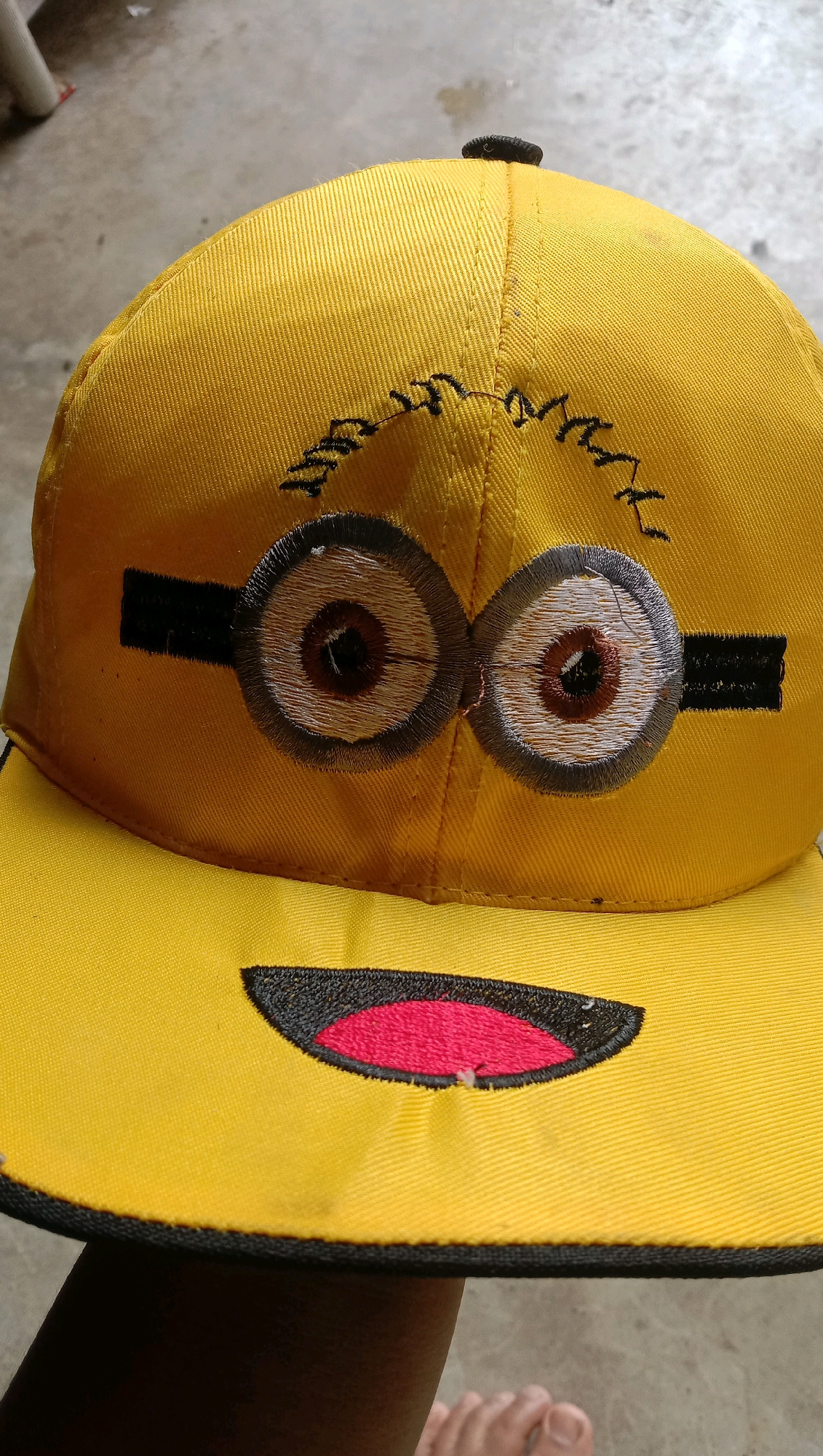 A yellow baseball cap, embroidered with the face of a Minion character from the Despicable Me franchise, is being held up by a person's hand and arm, partially obscuring the bottom edge of the cap. The Minion's face features large, circular eyes with brown irises and stitched black eyebrows above them. The cap also has a large, open mouth embroidered in red with a black outline. The background consists of a textured, concrete-like surface. The image was taken in the city of Kota Pari, Indonesia.