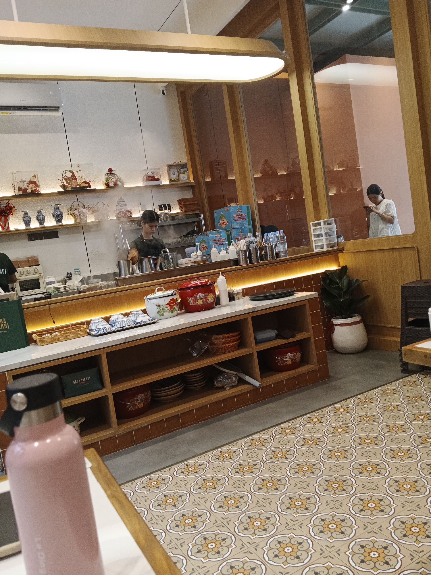 The image depicts the interior of a cafe in the city of Medan, Indonesia. The cafe features a counter area with a kitchen behind it, where a barista is preparing drinks. The counter is stocked with various items, including a red pot, a white pot with floral designs, and blue and white bowls. Shelves behind the counter display decorative items, vases, and packaged goods.
In the foreground, a pink water bottle rests on a table. The cafe's interior design includes wooden accents, warm lighting, and tiled flooring with a floral pattern. Through a glass partition, another woman is seen looking at her phone, while another person in the background is partially visible. The overall atmosphere suggests a bustling yet cozy dining establishment.