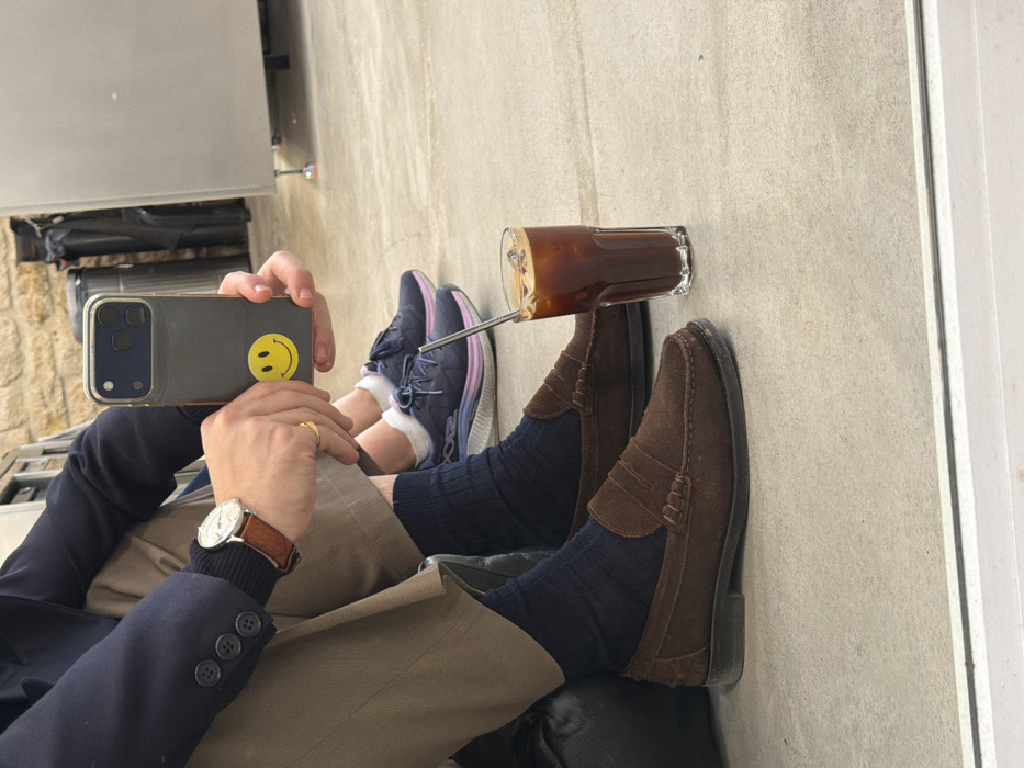 The image presents an indoor scene focused on the lower bodies of two individuals and a beverage. In the foreground, a person is seated, wearing light brown trousers, dark blue ribbed socks, and dark brown suede penny loafers. A silver-colored wristwatch with a brown leather strap is visible on their left wrist, and a gold ring is on their left ring finger. This individual holds a dark gray/blue smartphone, featuring a prominent yellow smiley face sticker on its back, as if taking a picture or video. Behind them, a second person's legs are visible, clad in white ankle socks and dark blue athletic shoes with lavender-pink soles. On the light gray tiled floor between the two individuals, a tall glass containing iced coffee with ice cubes and a silver metal straw is positioned. The background reveals a light-colored, possibly stone or textured wall, and industrial-style stainless steel equipment or furnishings to the right. The image depicts an indoor scene; however, insufficient visual information is present to determine a specific location within Paris, France.
