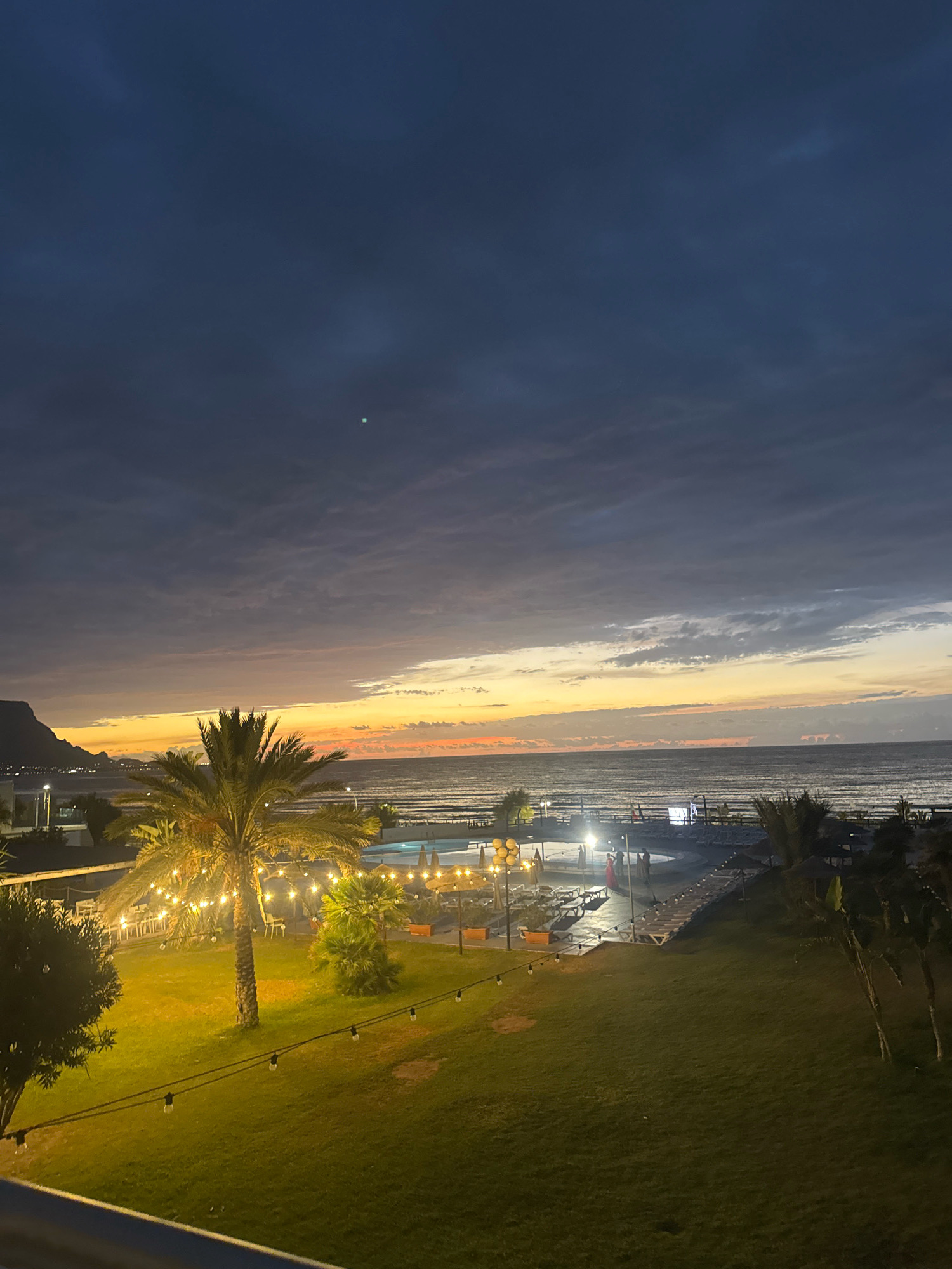 The image depicts a twilight scene at a resort or beachfront property in Isola delle Femmine, Italy. The sky is a deep blue transitioning to hues of orange and yellow near the horizon where the sea meets the sky. Clouds are visible, some appearing dark and heavy, others illuminated by the fading sunlight. A single, small, bright light, possibly a star or planet, is visible in the upper sky.
In the foreground, a lush green lawn is illuminated by string lights with visible bulbs. A prominent palm tree stands on the left side of the lawn, also adorned with string lights, casting a warm glow. Behind the palm tree and further into the scene, there is a swimming pool area with loungers and umbrellas. Other trees and foliage are scattered throughout the property.
Beyond the pool area, the sea stretches out, calm with visible ripples. The coastline is visible in the distance, with lights indicating buildings or a town. The overall atmosphere is serene and relaxing, suggesting an evening at a vacation destination.