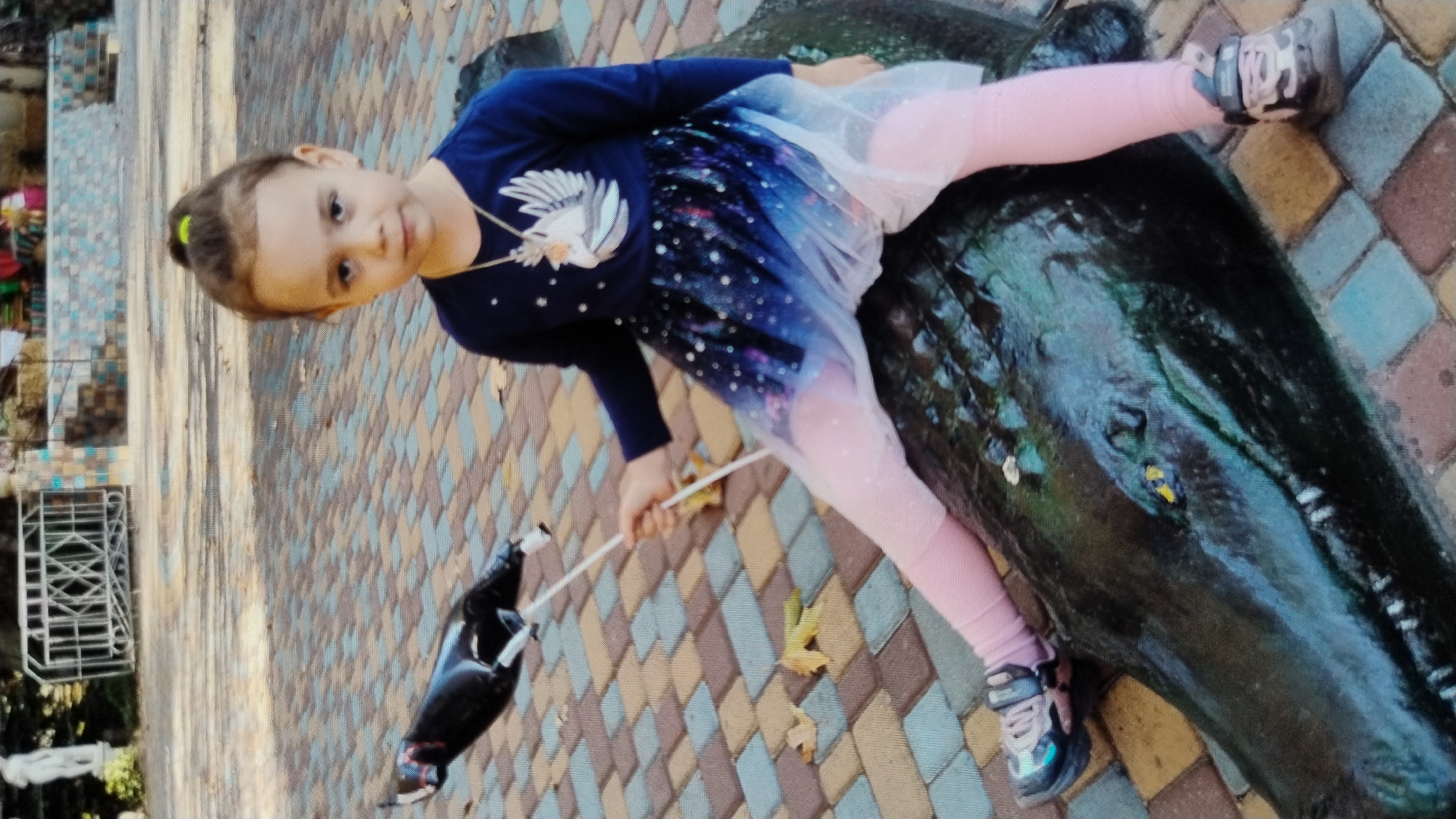 A young girl with brown hair pulled into a ponytail is sitting on a dark green, scaly sculpture that resembles a crocodile. She is wearing a dark blue long-sleeved shirt with a white bird emblem and a starry skirt that fades from dark blue to white. She also wears pink leggings and dark sneakers. In her right hand, she holds a white stick with a black balloon attached to it, which is partially obscured. The sculpture is placed on a paved area with multi-colored square tiles in shades of brown, beige, and blue. In the background, a white statue and a metal fence can be seen. The environment appears to be outdoors, possibly in a park or public space in Mykolaiv, Ukraine.