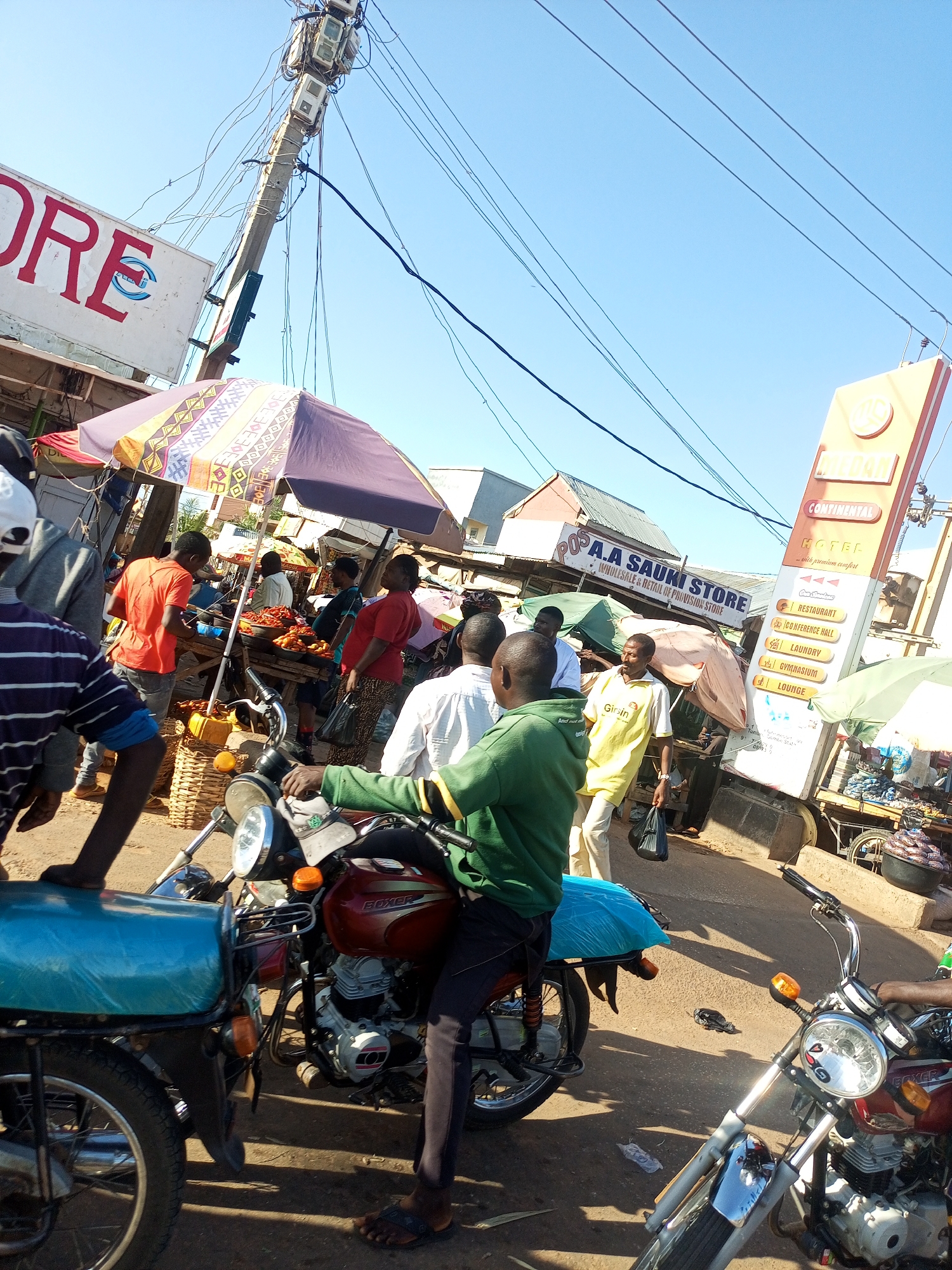 The image depicts a bustling street market scene in Tempure, Nigeria. In the foreground, two motorcycles are partially visible. The one on the left is blue and covered, while the one on the right is a red Boxer motorcycle with a person in a green jacket seated on it. The person's left hand is on the handlebars, and their right hand is not visible. A person wearing a blue and white striped shirt is standing to the left of the motorcycles.
In the background, several market stalls are set up under umbrellas. One stall has a sign that reads "A.A SAUKKI STORE WHOLESALE & RETAIL PROVISION STORE". A tall orange sign for "CONTINENTAL HOTEL" with various amenities listed, including "RESTAURANT", "CONFERENCE HALL", "LAUNDRY", "GYMNASIUM", and "LOUNGE", is prominently displayed on the right side of the image. Several people are engaged in various activities, such as selling goods and walking through the market. The environment appears to be a street with a clear blue sky overhead and overhead electrical wires. The ground is paved and appears dusty. The overall impression is one of a vibrant and active local marketplace.
