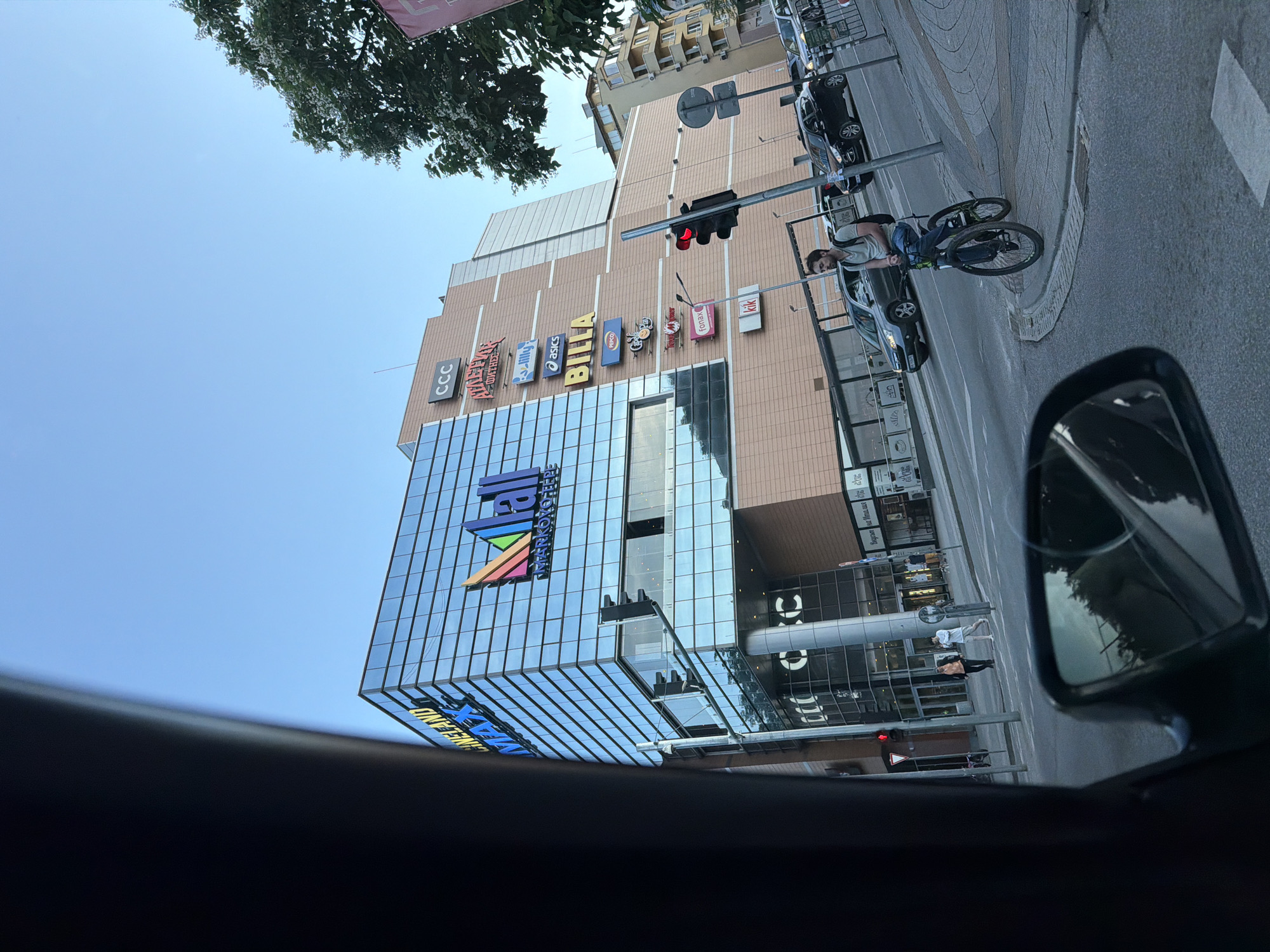 The image depicts a vibrant urban street scene in Plovdiv, Bulgaria, captured from the interior of a vehicle, looking towards a large commercial building. This multi-story structure, likely a shopping mall, features a modern glass facade on its lower sections, reflecting the clear blue sky, and a beige, brick-patterned exterior on its upper levels.
Numerous colorful brand logos are prominently displayed on the building's side, including CCC, LPP, Lilly Drogerie, Asics, Billa, Pepco, Deichmann, Fonal, and KiK. A large blue and rainbow-colored sign on the glass facade clearly reads "MARKOPOLO," with "CINEPALACE" and "KIDSLAND" visible below, identifying the building as "Mall Markopolo" which hosts a cinema and children's play area.
A paved street runs in front of the building, with several cars parked along the curb. A traffic light showing a red signal hangs over the street. To the right, a man wearing a backpack is stopped on a bicycle near the sidewalk, appearing to observe the street traffic. The viewpoint captures a portion of the car's dashboard in the foreground, and the side mirror on the right reflects a segment of the street behind, showing another pedestrian in the distance. Above, a clear blue sky is visible, along with the green leaves of a tree. The scene is brightly lit, indicative of daytime.