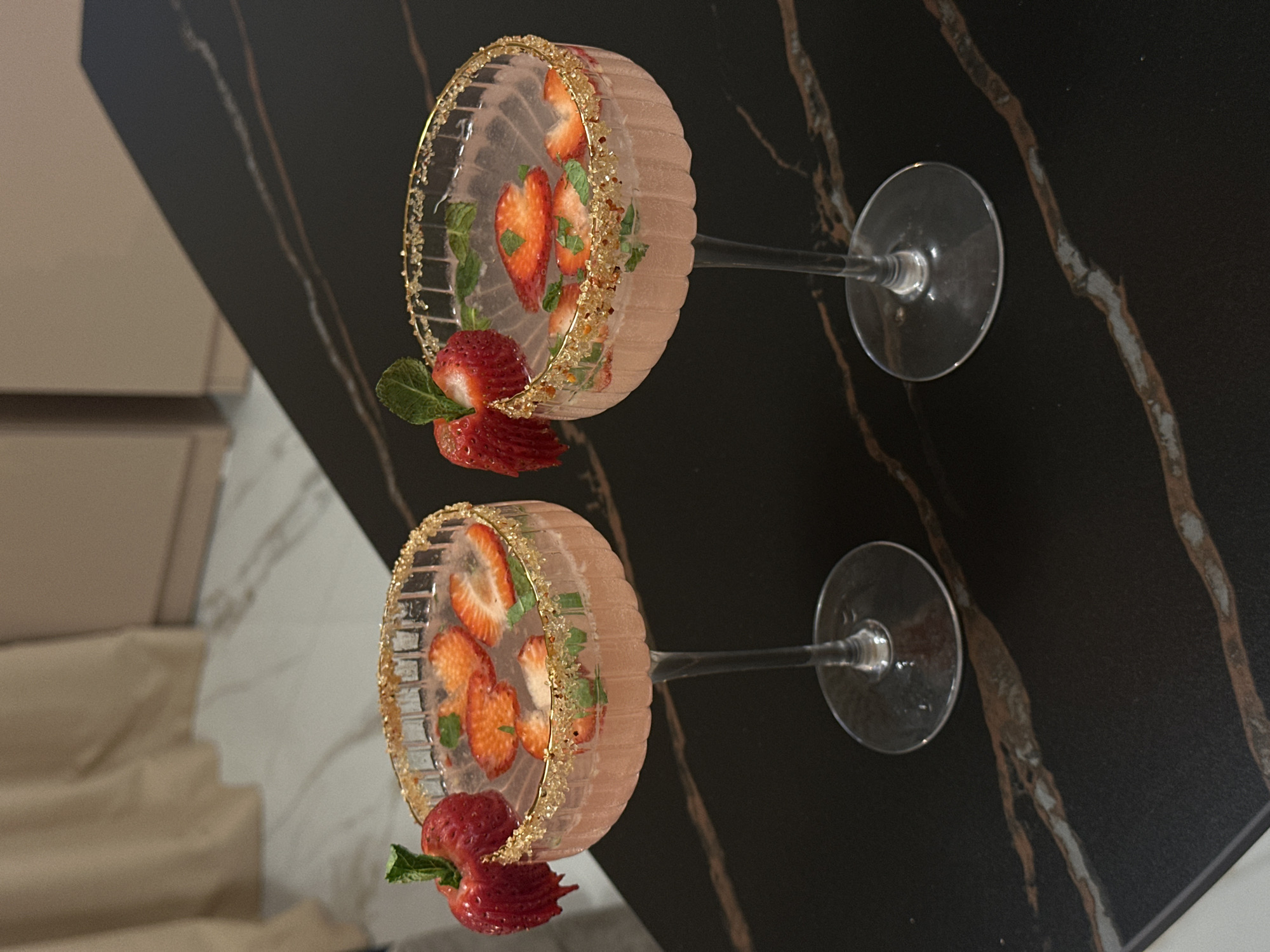 Two elegant cocktail glasses, possibly coupette or martini style, are displayed on a dark, veined countertop, likely marble or granite. The image is oriented sideways, presenting the glasses horizontally across the frame.
Each glass is filled with a light pink beverage, suggesting a strawberry-infused drink. The rims of both glasses are adorned with a generous coating of golden or bronze sprinkles, adding a festive touch. Inside the drinks, several slices of fresh strawberries and small green mint leaves are visible, floating amidst the liquid. Complementing the internal garnishes, a whole, fresh strawberry with its green stem and leaves is perched on the rim of each glass, slightly submerged in the drink. The bowls of the glasses feature a distinct ribbed texture.
The background is softly blurred, revealing light-colored furniture or cabinetry on the left and a more intricate, possibly marble, pattern on the right side behind the glasses. The overall setting appears to be indoors, possibly a kitchen or bar area, under artificial lighting. There are no people visible, and no discernible text appears in the image. The scene suggests a prepared presentation of refreshing drinks.