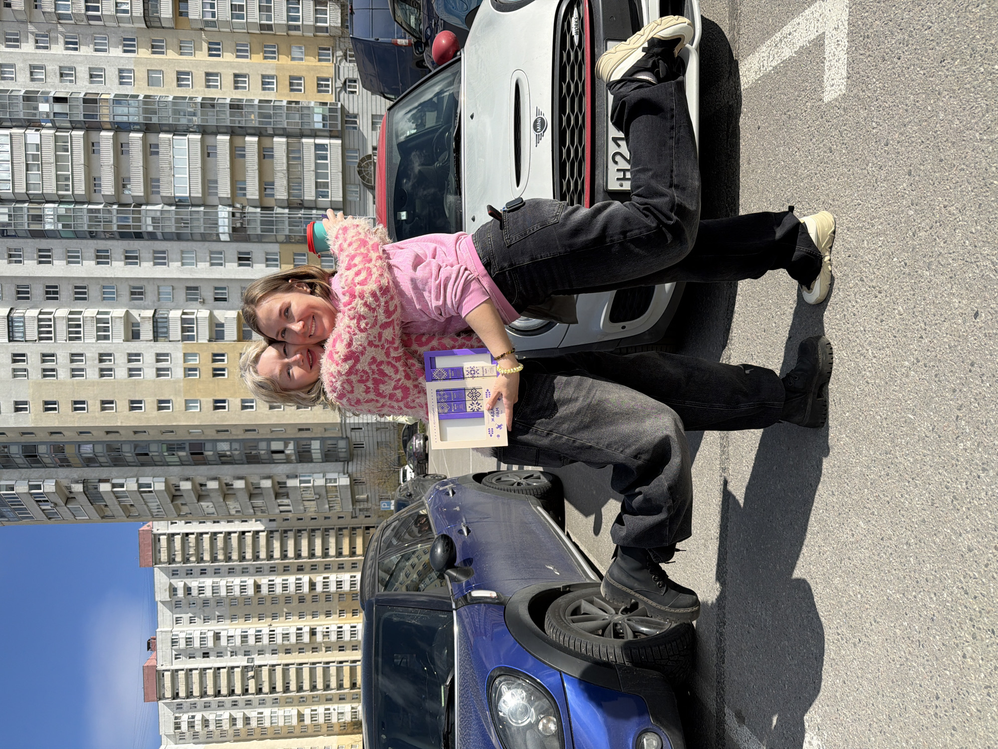 Two women are posing in front of two parked Mini Cooper cars. One woman is wearing a pink sweater and black jeans, and her legs are bent to pose on the hood of a white Mini Cooper. The other woman is wearing black jeans and is posing with one foot on the tire of a blue Mini Cooper. They are standing in a parking lot, with a tall apartment building visible in the background. The license plate on the white Mini Cooper is visible and reads "H21." Based on the architecture of the buildings, the location is likely in Saint Petersburg, Russia.