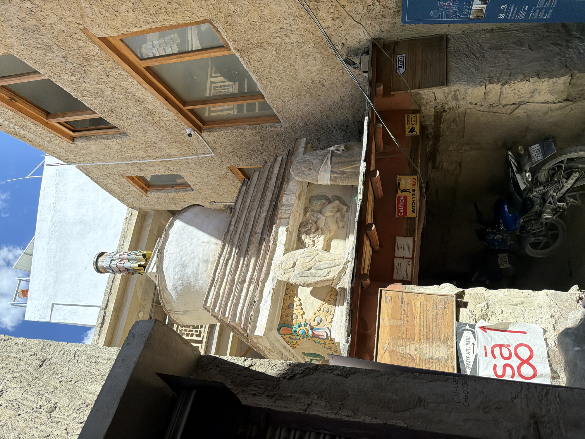 The image depicts a narrow alleyway in Leh, India, with traditional Ladakhi architecture. In the center, a white stupa, a Buddhist reliquary monument, is prominently displayed. It is adorned with colorful prayer flags and intricate carvings, including figures and floral motifs on its base. The stupa rests on a layered pedestal, which is supported by wooden beams.
Below the stupa, the alleyway narrows into a dimly lit passage. A sign indicating "SPINDLE ART STUDIO" with an arrow pointing forward is attached to a stone wall. To the right of this sign, a white banner with red lettering, "sā ∞", is visible. Further down the passage, a blue motorcycle is parked.
Above and to the right of the stupa, a beige, textured building with wooden-framed windows rises. A white building with solar panels and an antenna is visible in the background, suggesting a mix of traditional and modern elements. Several signs are affixed to the walls, including caution signs for low ceiling and a warning about CCTV operation. The sky above is blue with scattered clouds. The overall scene conveys a sense of cultural heritage and the everyday life of the city.
