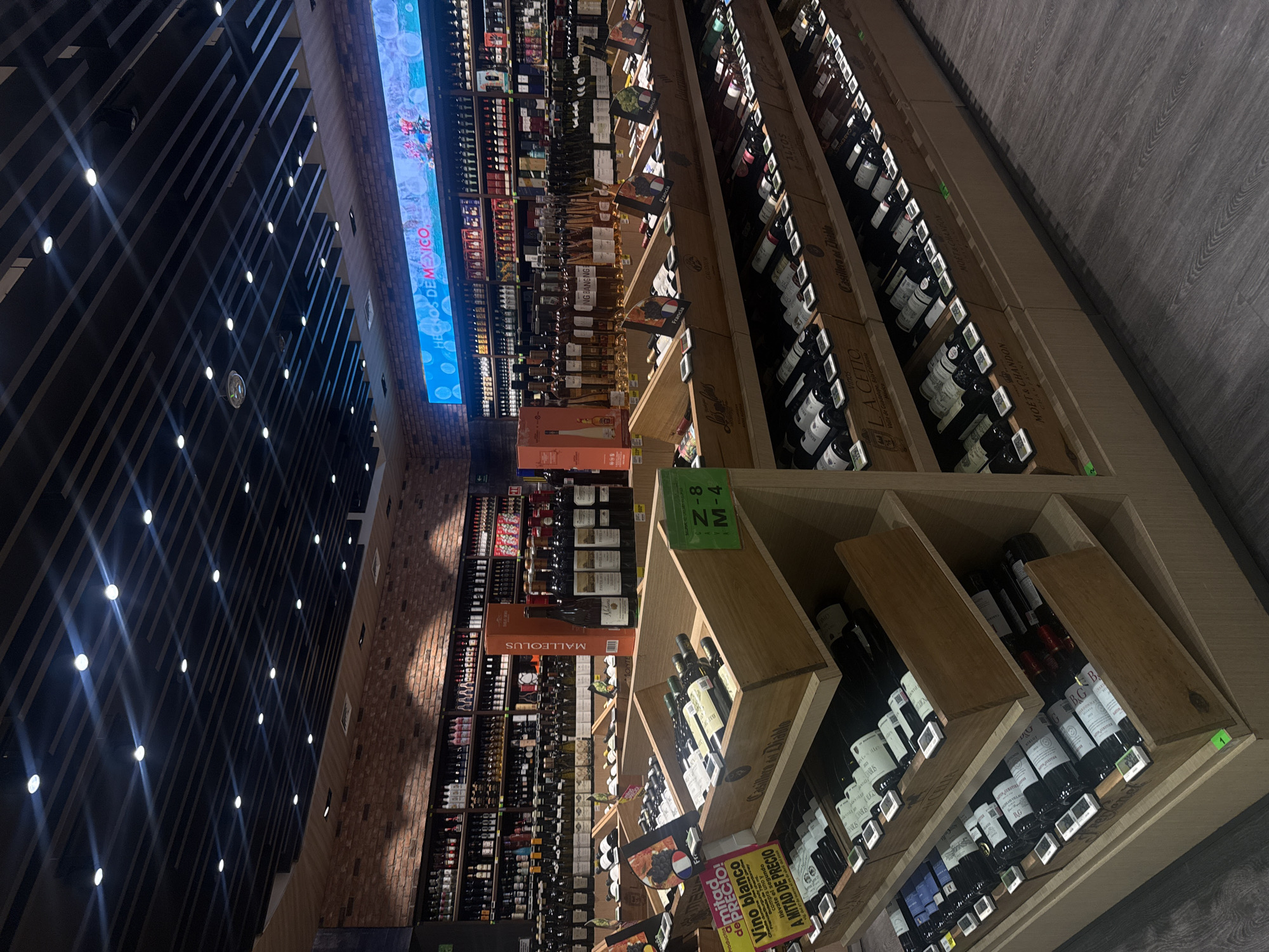 This indoor image shows a wine store. The scene is filled with rows of wine bottles arranged on wooden shelves. The bottles are of various brands and types. The shelves are stacked high, creating a vast selection of wines. The store is brightly lit, with a display screen showing "HECHOS DE MEXICO". Signage also includes "Z-8 M-4" and "MALLEOLUS". The environment includes a dark ceiling structure with many recessed lights, and a light-colored wood-grain floor.