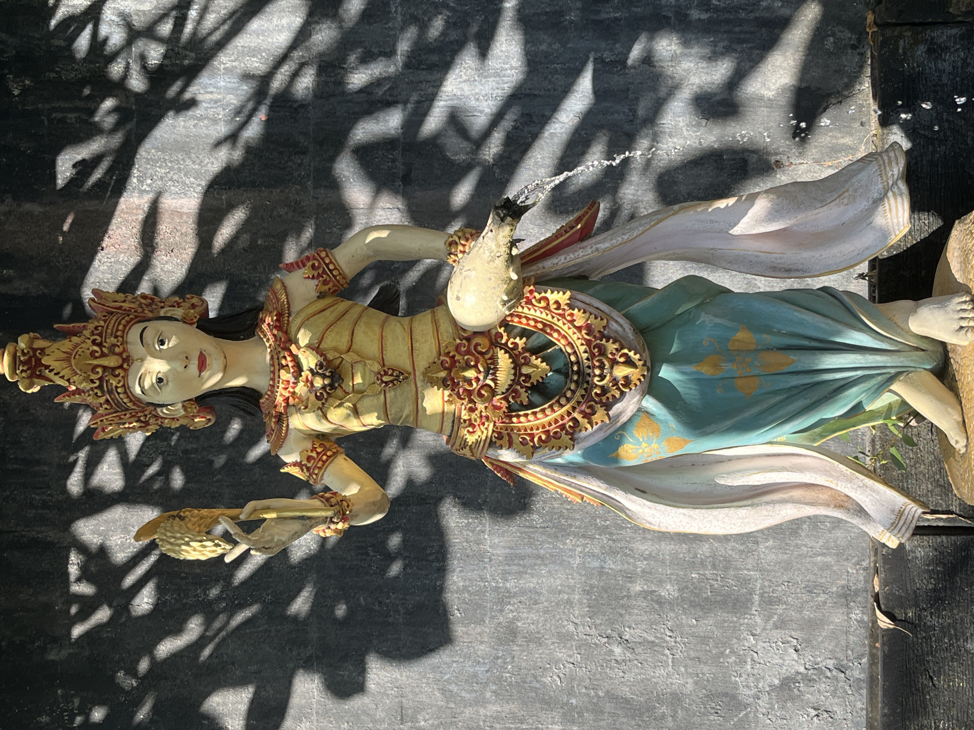 A full-shot statue of a female figure stands in front of a dark, textured wall. The statue is adorned in traditional attire with a golden crown, ornate jewelry, and a flowing skirt. She holds a golden object in her left hand and a white jug in her right, from which water is cascading. The wall behind her is partially obscured by dappled shadows cast by foliage. The statue is situated in Kuta Selatan, Indonesia.