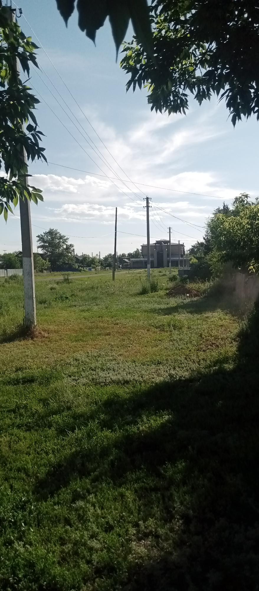 The image depicts a rural landscape scene in Marinovka, Kazakhstan, under a bright, partly cloudy sky suggesting it is daytime. The foreground is dominated by a grassy field with a tall, light-colored utility pole on the left side, partially obscured by foliage from an overhanging tree. Several other utility poles with wires are visible further into the scene, stretching across the sky.
In the mid-ground, a partially constructed building, appearing to be a house or a larger structure with multiple levels and visible concrete walls, stands on the right side of a dirt road. This building is surrounded by greenery, with trees and bushes prevalent throughout the area. Various other smaller structures, possibly houses or outbuildings, are scattered in the distance, indicating a residential or semi-rural area.
The overall atmosphere is one of a quiet, possibly developing, rural community on a clear day. There is no visible text in the image, nor any people or specific events taking place. The scene is characterized by natural elements like grass, trees, and sky, interspersed with man-made structures and utility infrastructure. The lighting suggests a sunny day, likely late morning or afternoon, with shadows cast on the grass.