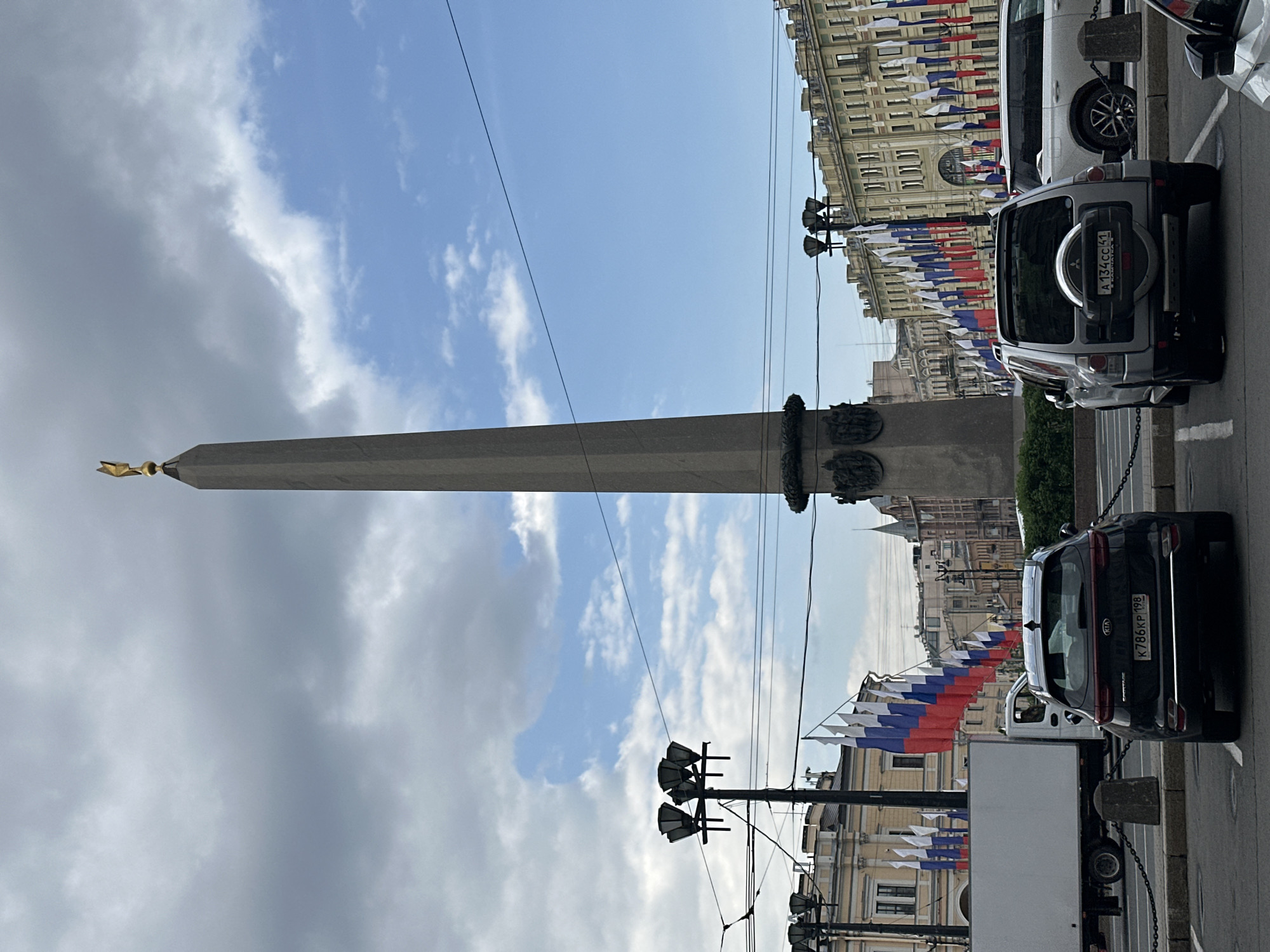The image captures a street scene in Saint Petersburg, Russia, featuring the Alexander Column prominently. The column is topped with a golden angel figure and stands tall against a backdrop of a partly cloudy blue sky. Below the column, the foreground is occupied by parked cars, including a silver Mitsubishi with the license plate "A 134 CC 47" and a dark-colored Kia with the license plate "K 786 KP 198." Buildings line the street, adorned with Russian flags. Streetlights and overhead wires add to the urban setting.
