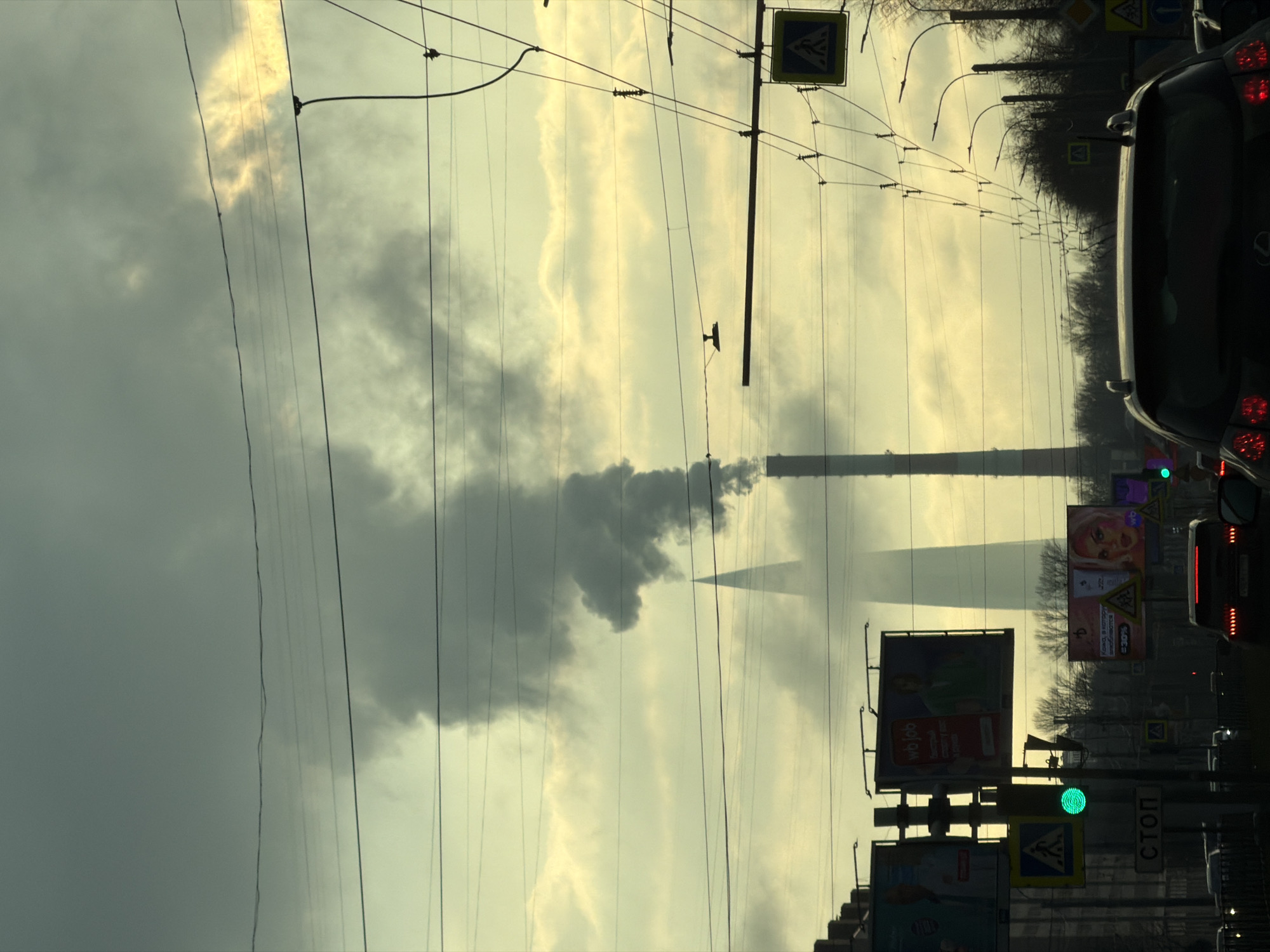 The image captures a street scene in Saint Petersburg, Russia, characterized by overcast skies and industrial elements. A tall chimney stack, likely part of a power plant, is visible emitting smoke into the air. Overhead power lines crisscross the sky. A line of billboards and traffic lights line the street. A dark-colored vehicle is positioned in the right of the frame. Pedestrian crossing signs are seen along the roadside.