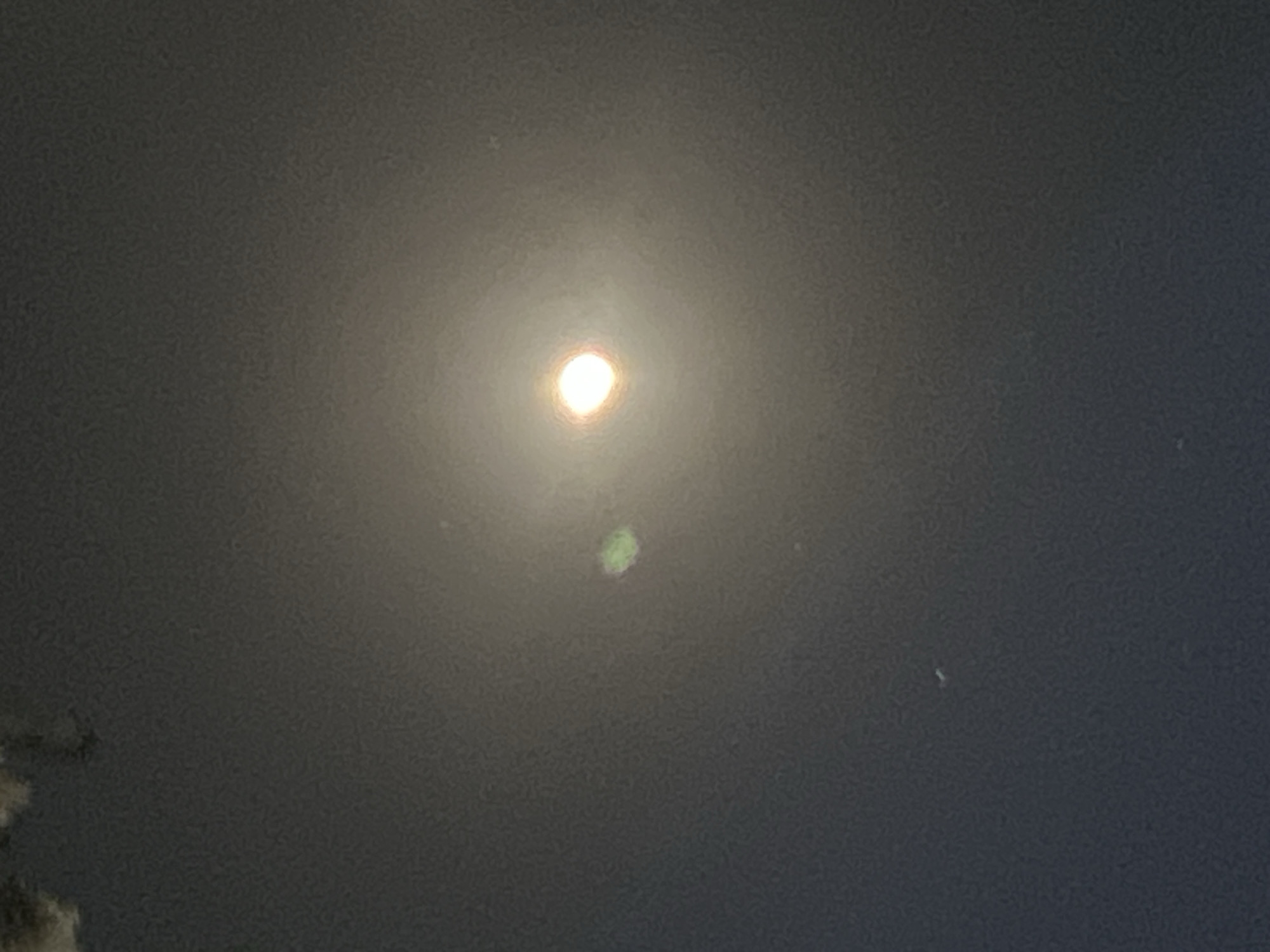 The image depicts a nighttime sky with a brightly lit object, likely the moon, as its central focus. There are some indistinct cloud formations in the upper left portion of the image. A faint green circular anomaly is visible to the left of the bright object. The overall ambiance is dark with a soft, diffused light emanating from the central celestial body. There are no visible indications of the city of Denpasar, Indonesia, or any human presence within the frame.