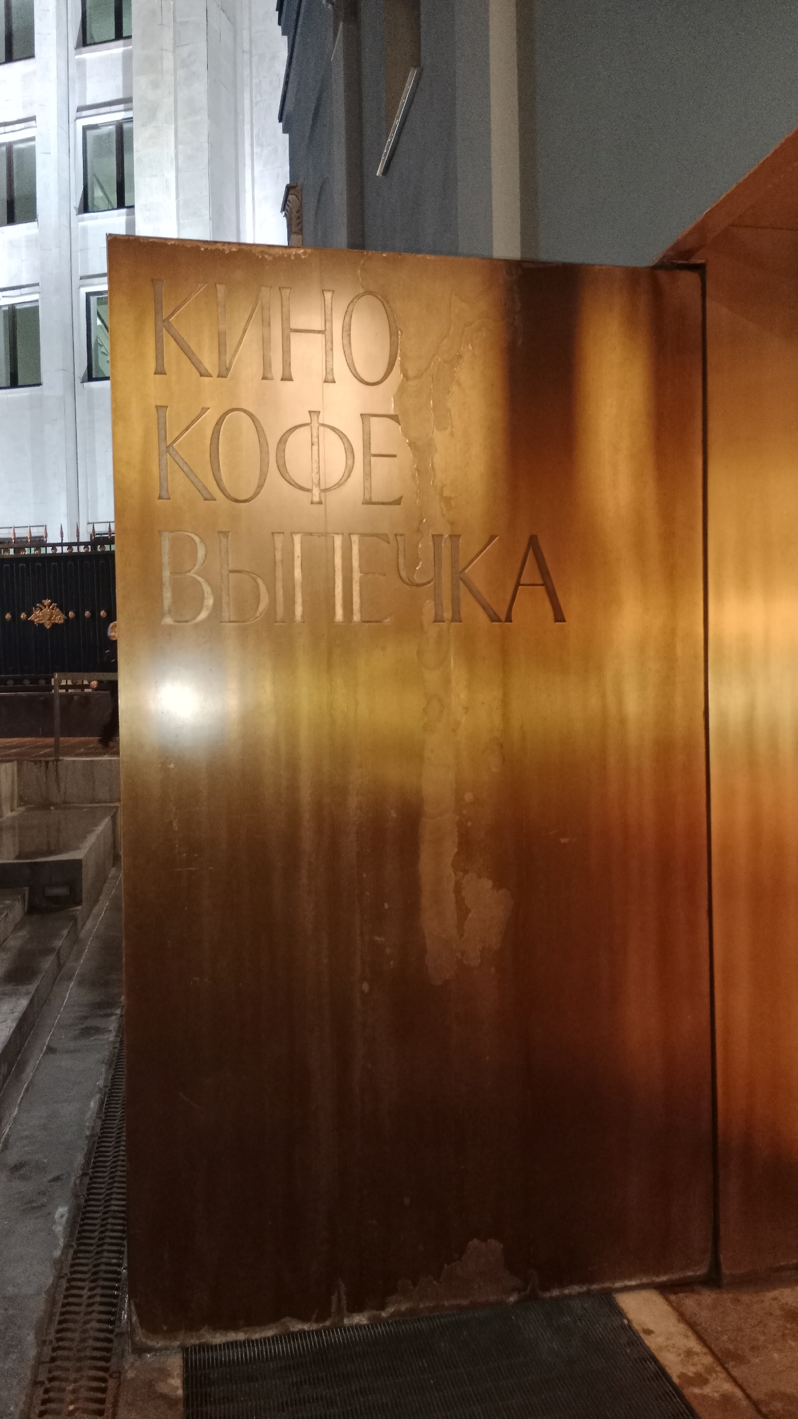 A close-up shot captures a bronze-colored sign in Moscow, Russia. The sign features inscriptions in Russian: "КИНО," "КОФЕ," and "ВЫПЕЧКА," which translate to "Cinema," "Coffee," and "Pastries." The sign is affixed to a building or structure, likely indicating the presence of these services nearby. The texture of the sign shows signs of weathering and moisture stains. The background includes glimpses of a building facade with windows and a metal fence. The lighting suggests an evening or indoor setting.