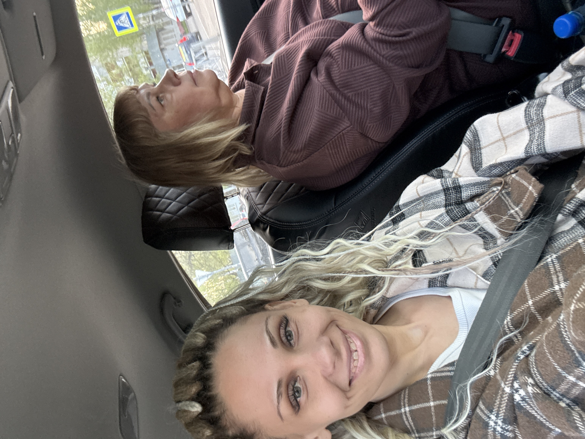 Two females are seated in a car, presumably in Rusanovo, Russia. The foreground woman, with blonde braided hair, smiles at the camera. She wears a white tank top under a brown and white plaid shirt, secured by a seatbelt. Behind her, another woman with short blonde hair is looking out the window. She wears a brown top and is also secured by a seatbelt. Through the car window, a street sign is visible. The car's interior is dark, with leather seats.