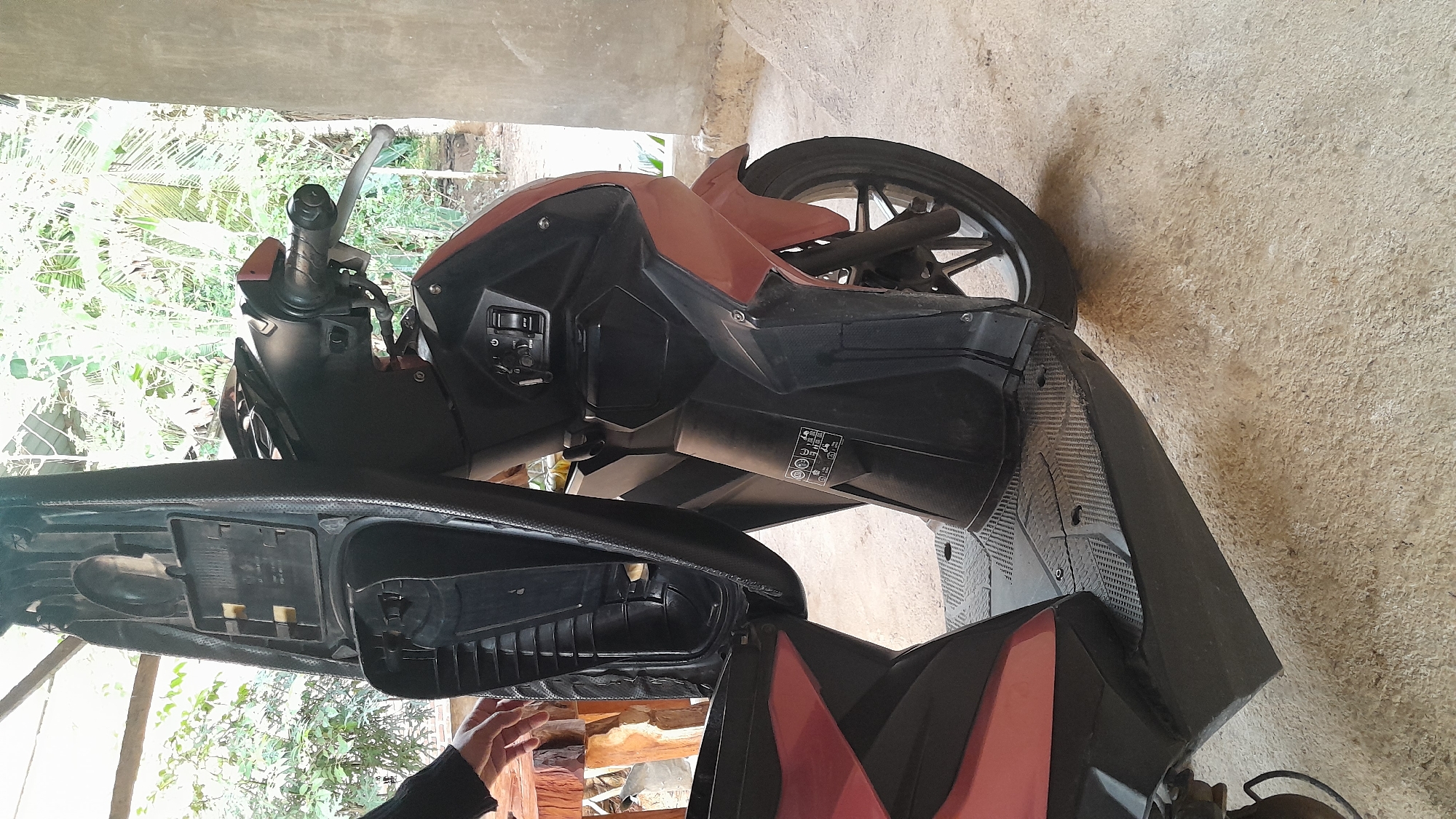 The image shows a parked scooter in Aluerambee, Indonesia. The scooter is predominantly black with some reddish-brown accents. Its seat is open, revealing the storage compartment. The environment appears to be a covered outdoor area, possibly a garage or shed, with a view of foliage and trees. The ground is a light-colored concrete surface. A person's hand is visible near the scooter's open compartment.