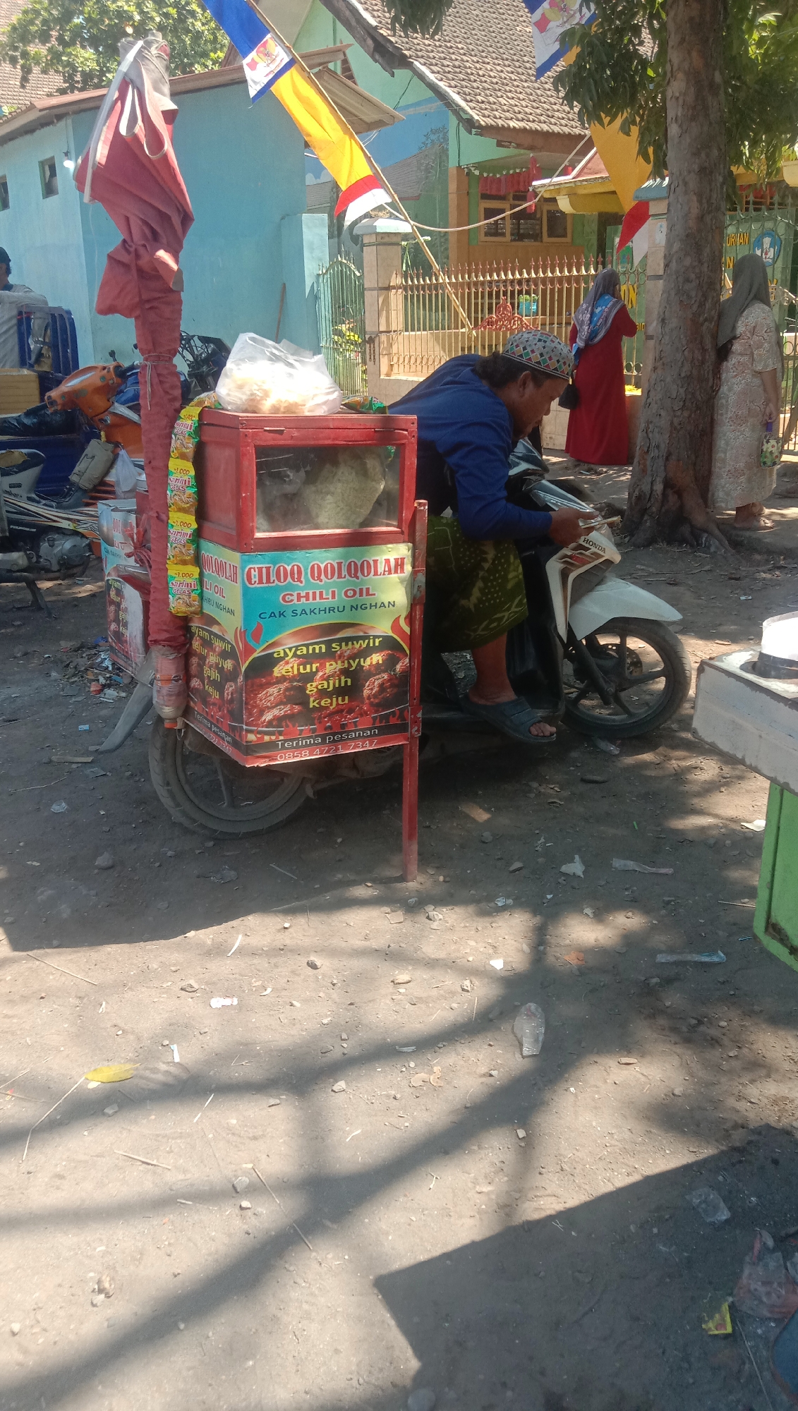 The image captures a street scene in Ngempit, Indonesia, with a focus on a food cart. A person, identified as a male wearing a blue shirt and a patterned headscarf, is seated on a white Honda motorcycle. The motorcycle is parked behind a red food cart, which is adorned with colorful graphics advertising food items. The cart has a glass display case on top, containing what appears to be food items, and the side of the cart displays text in Indonesian, including "CHILI OIL," "AYAM SUWIR," "TELUR PUYUH," "GAJIH," and "KEJU." The cart also has a phone number displayed for orders. In the background, there are buildings with tiled roofs and a fence. Two women, one in a red dress and the other in a patterned dress, are visible near a tree. The ground is unpaved and dusty, with scattered debris, and shadows from trees are cast across the scene.