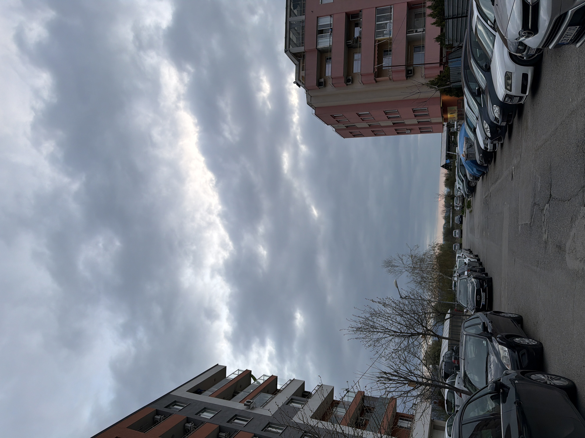 The image shows a street scene in Sofia, Bulgaria, with buildings on either side and cars parked along the street. The sky is overcast with grey clouds. A pink building is on one side and a grey building with orange-brown accents is on the other. The street is paved and appears to be slightly cracked or uneven. Bare tree branches are visible along the street, in front of the grey building. Cars of various makes and colors are parked in a row, mostly facing in the same direction. The lighting suggests it is either early morning, late afternoon, or a cloudy day.