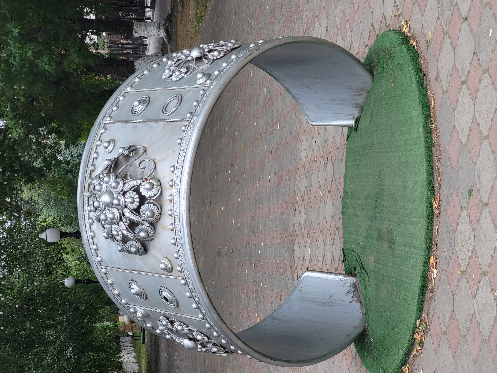 This is a medium, eye-level, full shot of a large, silver, horseshoe-shaped sculpture in a park. The sculpture is decorated with raised, circular elements and floral motifs. It sits on a circular patch of artificial green grass. The ground around the grass is paved with a diamond pattern of red and beige bricks. In the background, there are trees with green foliage, two lampposts, and a dark metal fence. The lighting is overcast. The sculpture appears to be located in the city of Petropavl, Kazakhstan.