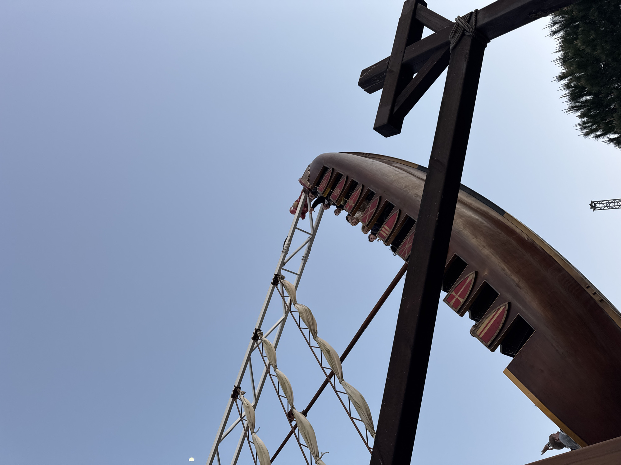 The image captures a "Viking Ship" amusement ride in Sochi, Russia. The ride, shaped like a Viking ship, is brown with red and yellow shield decorations along the sides. Several riders are visible within the ship's seating area. The ship is attached to a white metal frame supported by ropes. A dark brown wooden structure with a cross-shaped beam is adjacent to the ride. The background consists of a clear blue sky, with a small section of green foliage visible in the upper right corner. A construction crane is also faintly visible in the background. The perspective is from a low angle, looking up at the ride.
