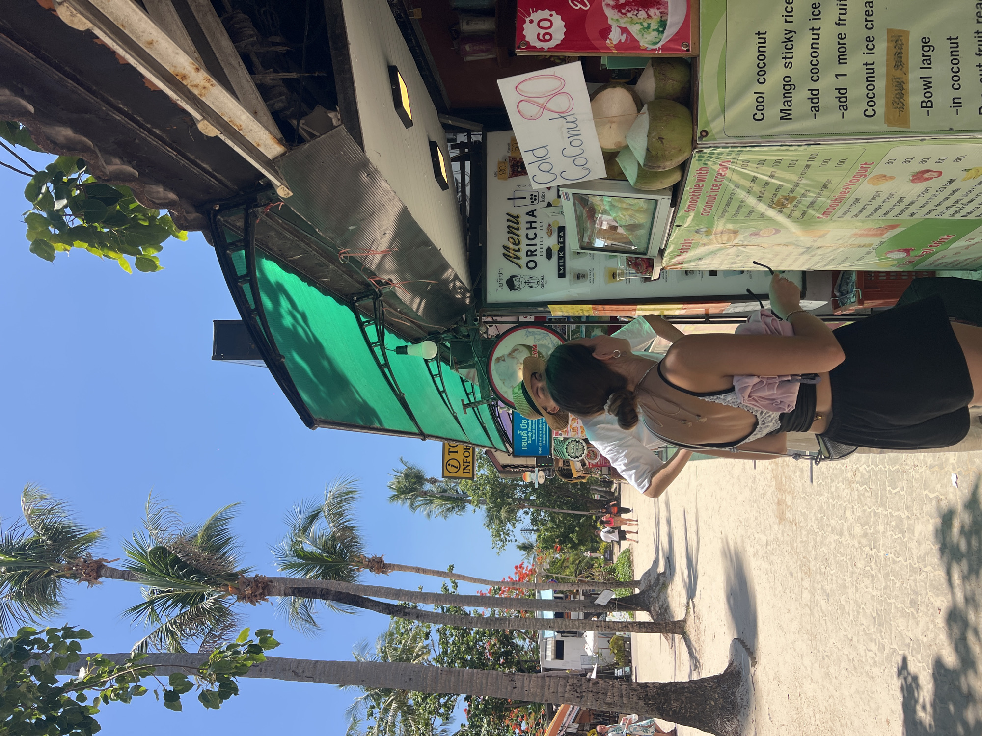 A tourist in Ban Ao Nang, Thailand, is looking at a menu on a street vendor stall selling smoothies, coconut ice cream, and cold coconuts. The menu is written in both Thai and English. The stall also sells Oicha bubble tea. The menu displays the prices for each item. The stall is located on a street lined with palm trees. Other people can be seen walking in the background.