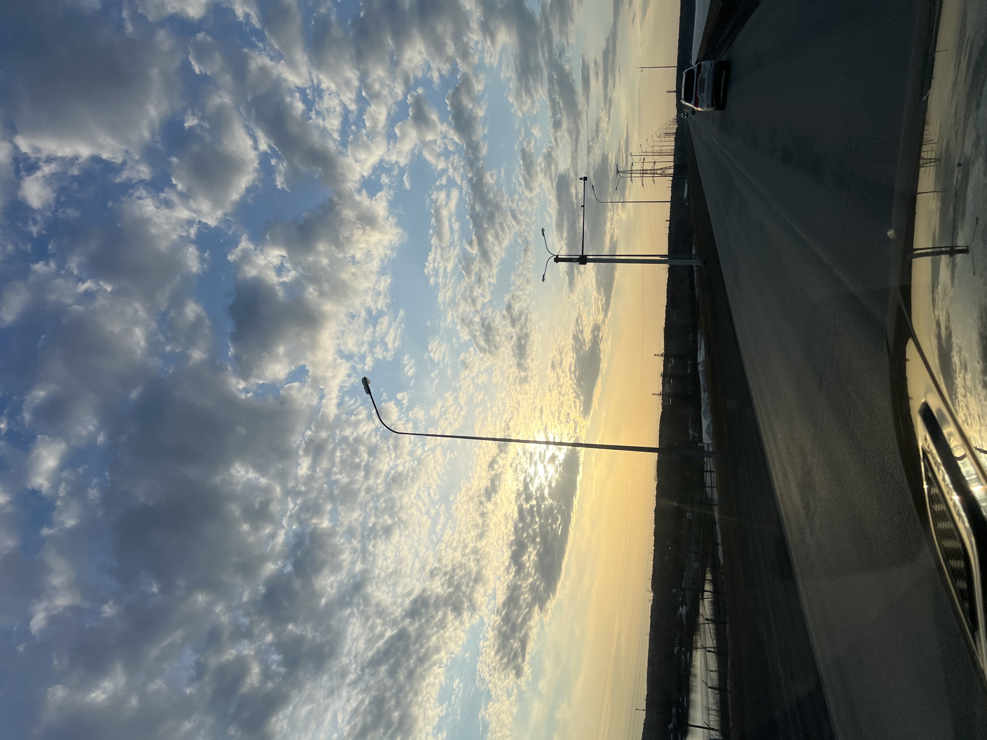 A roadside view in what appears to be Chaikovsky, Russia. The photograph, taken from inside a car, captures a straight, black asphalt road illuminated by the setting sun. A single car is visible on the road. Numerous street lights line the road's shoulder. Beyond the road, a landscape of low shrubs or trees is visible. The sky above is a mix of bright blue and white clouds tinged with the golden hue of the setting sun. The sky's reflection is also visible on the wet surface of the road.