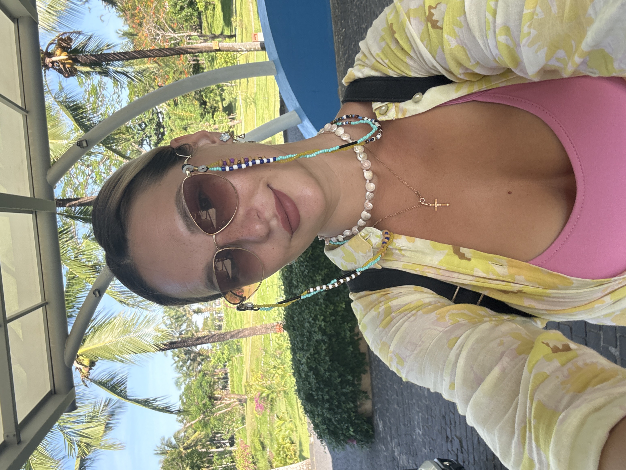 A woman is taking a selfie in front of a building with palm trees and lush greenery in the background. She is wearing sunglasses, a yellow and white patterned shirt, and a pink top. A beaded chain is attached to her sunglasses and rests on her chest, where she is also wearing necklaces. The location appears to be outdoors, possibly a resort or hotel in Đà Nẵng, Vietnam, given the presence of tropical vegetation and modern architecture.
