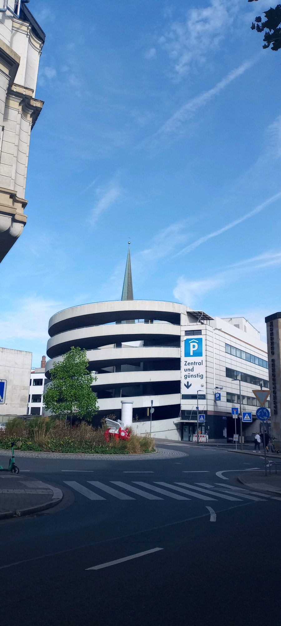 The image shows a modern, circular, multi-level parking garage in Dortmund, Germany. The garage has a distinctive spiral ramp design. A tall, green spire, likely belonging to a church or other public building, rises from behind the garage. The sky is blue with wispy clouds and a contrail. In the foreground, there is a street intersection with a pedestrian crossing. A green electric scooter is parked on the left side of the street. In the center of the roundabout, there is a small landscaped island with grasses, plants, and a whimsical red sculpture of a winged pig. Signs on the parking garage indicate "Zentral und Günstig" (Central and Affordable) with a parking symbol. Several pedestrians are visible walking on the sidewalk near the entrance to the parking garage. The overall scene is a typical urban environment.