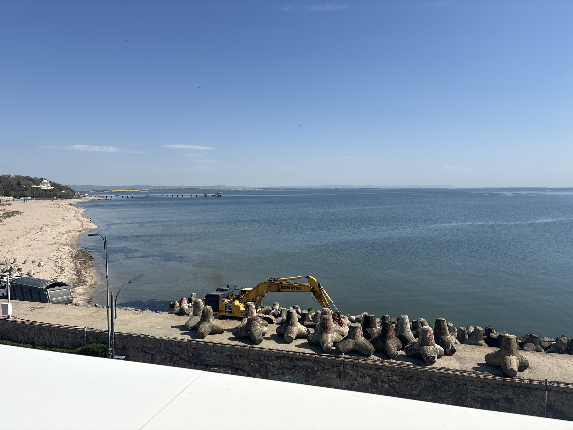 This wide-angle image captures a sunny coastal scene in Burgas, Bulgaria, featuring a beach, sea, and active construction work. The foreground is dominated by a white, flat surface, likely a balcony or building edge, beneath which a sturdy stone and concrete seawall is visible.
On the concrete promenade above the seawall, numerous grey, four-legged concrete tetrapods are systematically arranged. Many of these tetrapods bear faint red markings. A large yellow "KOMATSU" excavator is positioned among these units, suggesting ongoing work related to coastal defense or infrastructure development. To the left of the excavator, further along the promenade, stands a grey street lamp with two fixtures, and a large, dark, arched container or trailer is parked closer to the beach.
The sandy beach stretches along the left side of the frame, curving gently into the calm, clear blue-green sea. A few small human figures can be seen on the beach, enjoying the sunny weather. In the distance, a long pier extends into the sea from the beach, and beyond that, the coastline is dotted with distant buildings and green hills. A prominent white building with a distinctive roof sits atop a verdant hill on the far left. The horizon reveals a clear blue sky with only a few wispy clouds. The bright, even lighting indicates it is daytime, likely mid-morning or early afternoon. The overall impression is one of calm natural beauty coexisting with active coastal engineering.