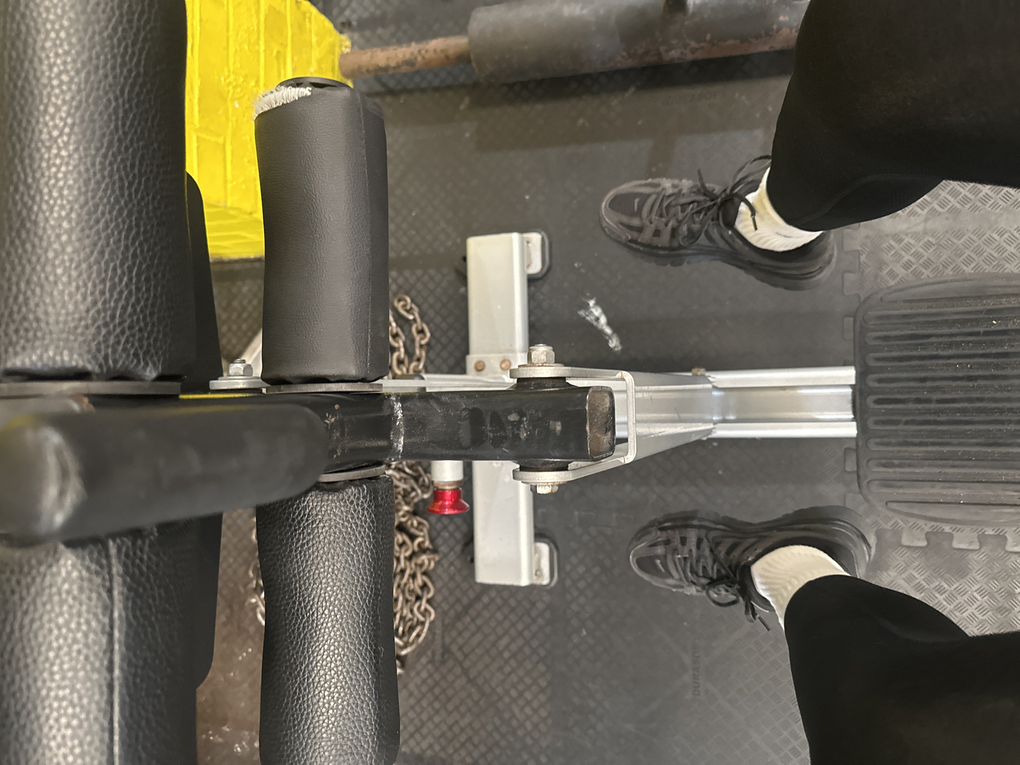 The image depicts an overhead view of a seated cable row machine, likely located in a gym, with a person positioned to use it. The machine's black padded seat and leg supports are visible, along with the silver cable attachment point and chain. The person's feet, clad in grey sneakers and white socks, rest on the footrests. The gym floor is covered in interlocking rubber mats. The location, Tunbridge Wells, United Kingdom, is not discernible from the visual information provided.