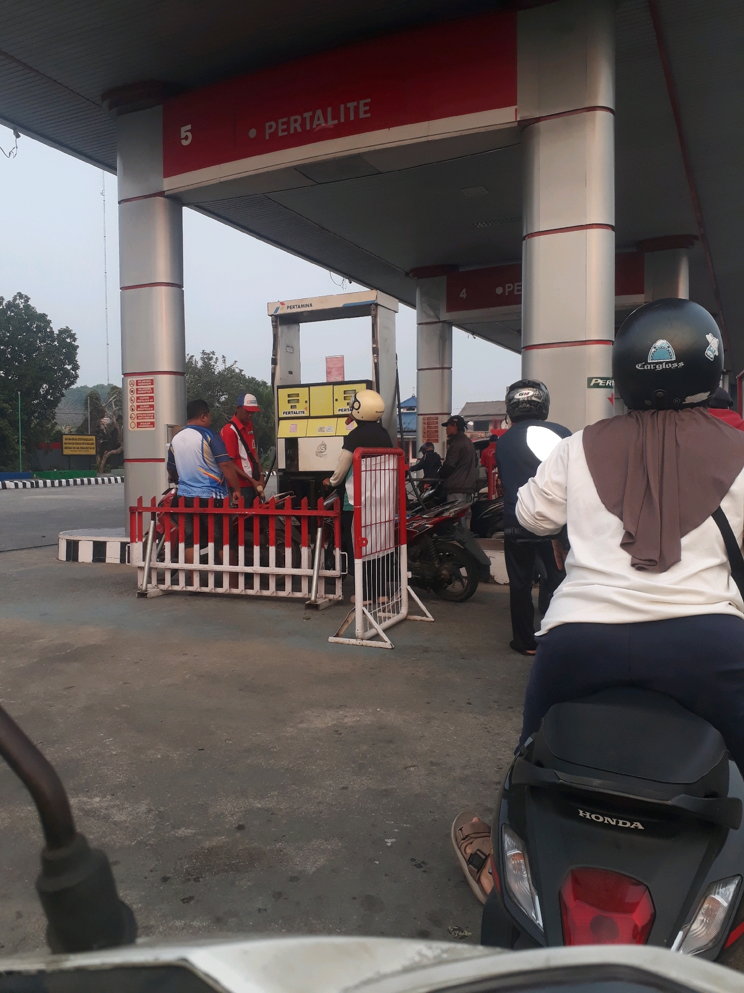 This image shows a busy scene at a Pertamina gas station in Pangkalpinang, Indonesia. Multiple people on motorcycles are waiting to refuel. A gas pump, labeled "PERTALITE," is visible, with a worker attending to a customer. The station has distinctive red and silver pillars supporting a canopy. The overall atmosphere suggests a typical day at a gas station, with vehicles and people engaged in the act of refueling.