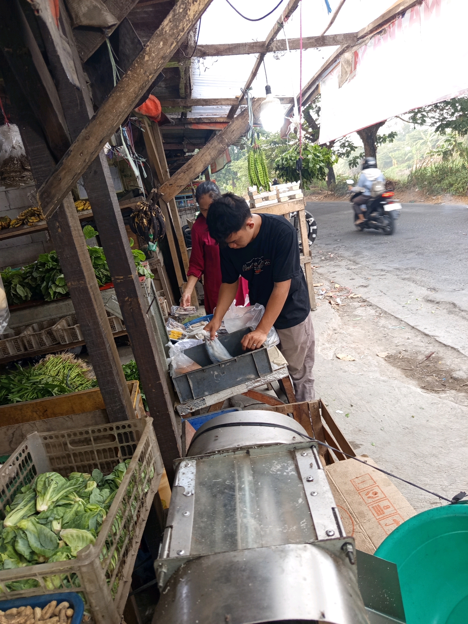 A man wearing a black t-shirt and tan pants is seen at an outdoor market stall in Karawang, Indonesia. He is reaching into a gray plastic crate, holding a plastic bag containing a fish. To his left, a woman in a red shirt stands behind the counter, holding a clear plastic bag filled with what appears to be food items. In front of them are crates of fresh vegetables, including leafy greens. Behind the stall, a road is visible with a motorcyclist passing by. The stall itself is constructed from rough wooden beams and covered with corrugated plastic sheeting. The overall scene suggests a busy street-side market.