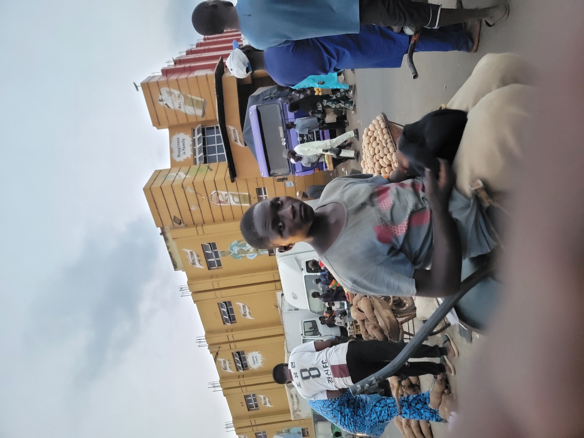 The image, taken in Jos, Nigeria, shows a street scene with a mix of commercial and human activity. A multi-story building, or series of stacked structures, dominates the left side of the frame, covered with "MALTINA" branding, suggesting an advertisement or product placement. In the foreground and middle, several individuals are visible, including a person looking directly at the camera. There's a variety of produce displayed, including what appears to be eggs and yams. Vehicles, including a truck, are visible further down the street. People are scattered around, some standing, some in transit. The sky is overcast, indicating possible weather conditions. Overall, the scene captures a busy, informal market environment.