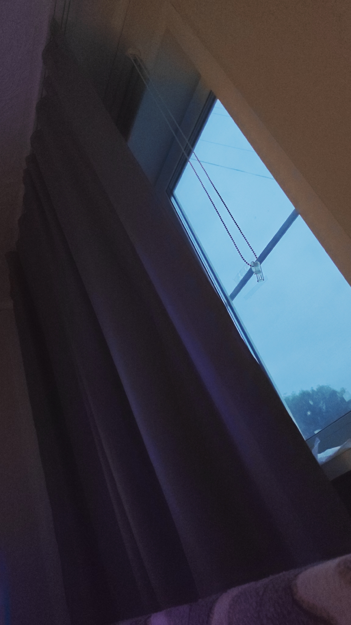 The image displays a window with a blue tint, likely from an overlay or the sky. A beaded chain hangs from the top of the window frame, attached to a small white mechanism. To the left of the window, dark purple curtains are drawn. In the foreground, a blurry surface with purple and cream stripes is visible, possibly a blanket or upholstery. The overall scene is dimly lit, with the bright window providing the primary light source. The image is taken from a low angle, looking upwards towards the window. There is no indication of specific location within Redkino, Russia, beyond the fact that the image is presented in that context.