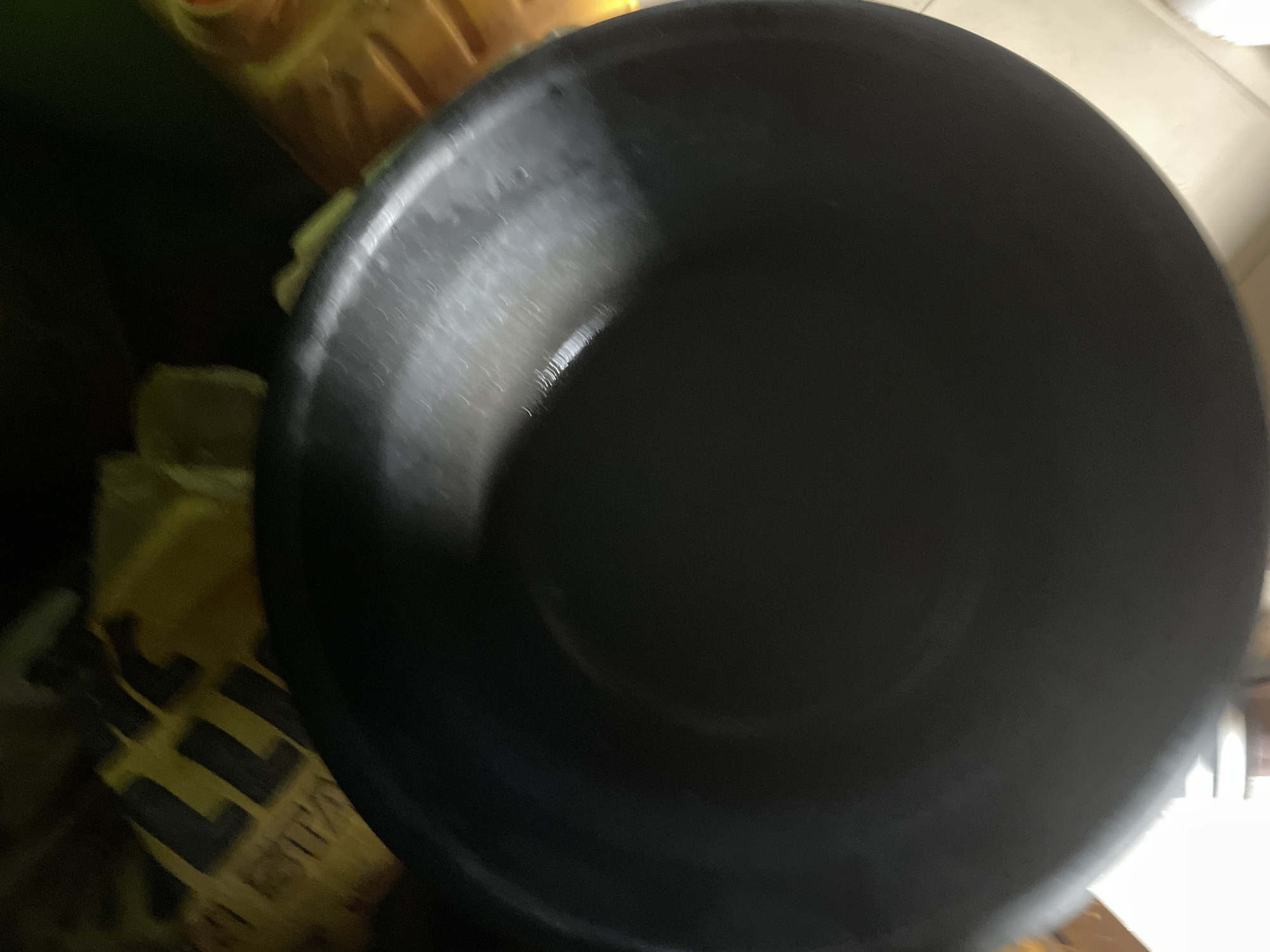 The image shows a close-up of a black, shallow, round object, possibly a bowl or basin, with some reflective light on its surface. In the background, there are blurry yellow objects with black lettering, which appear to be some form of packaging or containers, and a dark green background. The scene is dimly lit. There are no people visible in the image. The location in Jalingo, Nigeria, is not identifiable from the visual information provided in the image.