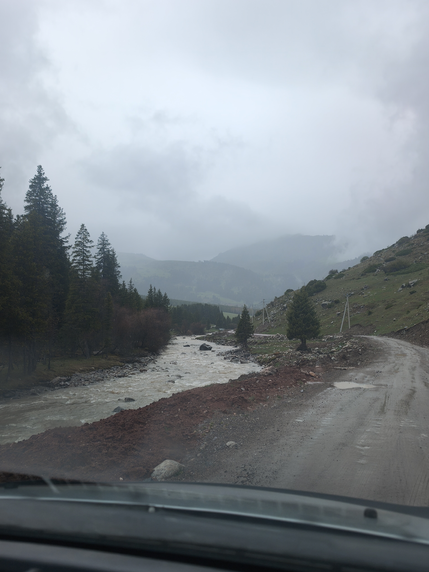The image presents a mountainous landscape in Dzhetyoguz, Kyrgyzstan, captured from the interior of a vehicle, likely through its windshield. The dominant feature is a heavy, overcast sky, with thick grey clouds obscuring the tops of distant mountains and creating a muted, diffused light.

In the foreground, a wet, unpaved dirt road with several puddles curves to the right, indicating recent rainfall. To the immediate left of the road, a significant pile of reddish-brown earth and rocks lines the bank of a fast-flowing, brownish-grey river. The river itself is turbulent and wide, likely swollen by rain or snowmelt, and its banks are strewn with rocks and smaller debris.

Along the left side of the frame, a dense line of tall coniferous trees, possibly spruce or fir, stands beside the river, their dark green foliage creating a stark contrast with the cloudy sky. Further into the mid-ground, the landscape opens to reveal rolling hills and more mountains partially obscured by mist and low-hanging clouds. The right side of the frame shows a grassy hillside dotted with rocks and occasional smaller trees, with several utility poles running parallel to the road, suggesting a power line or communication infrastructure.

The overall scene conveys a sense of wildness and remoteness. The weather appears to be overcast and possibly damp, contributing to a serene yet somber atmosphere. No people or visible text are present in the image. The time of day is daytime, but the heavy cloud cover prevents bright sunlight.