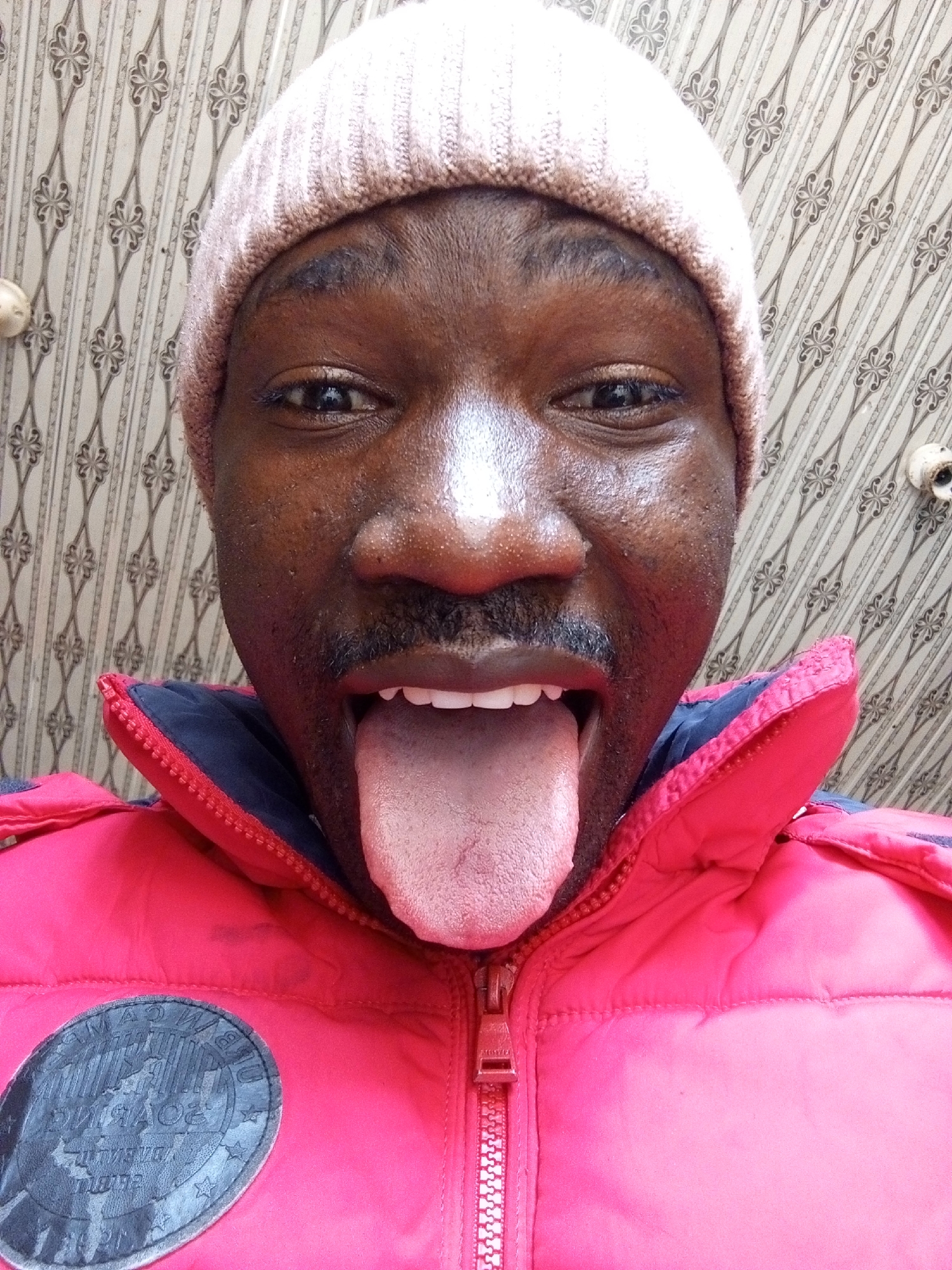 A close-up selfie shows a dark-skinned man wearing a pink beanie and a red puffy jacket with a dark gray circular patch on the left side. He is sticking his tongue out, and the background appears to be a patterned wallpaper with off-white and gray tones. No contextual details indicate a specific location within Gwong, Nigeria.
