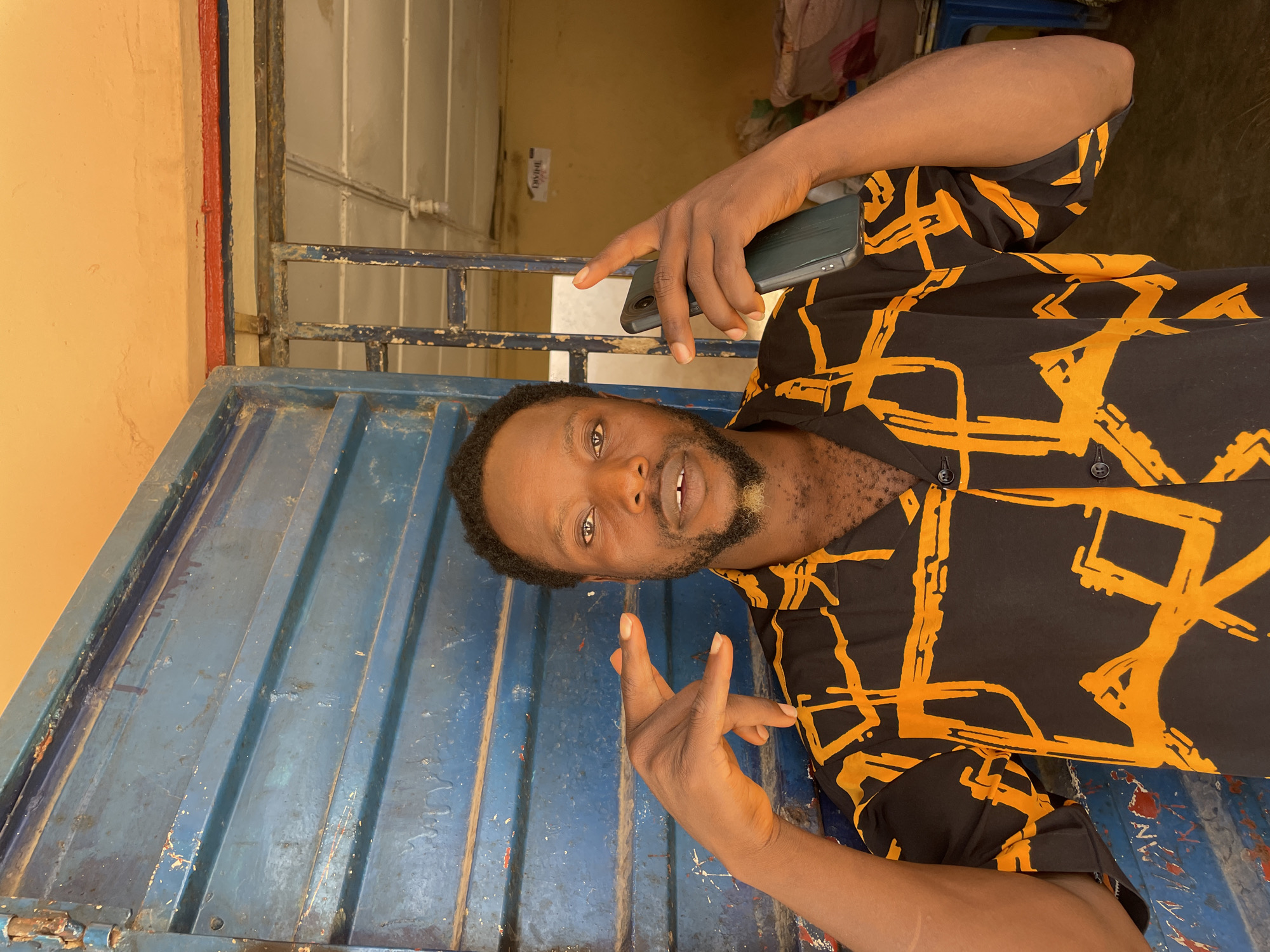 A man with dark skin and a beard, wearing a black shirt with an orange abstract pattern, poses with his hands in a gesture in front of a blue, ribbed metal door. He holds a dark gray smartphone in his right hand. The background appears to be an interior space with yellow walls and a barred window. A sign with the word "DIVINE" is visible in the distance. The scene is likely in Jos, Nigeria, given the context.