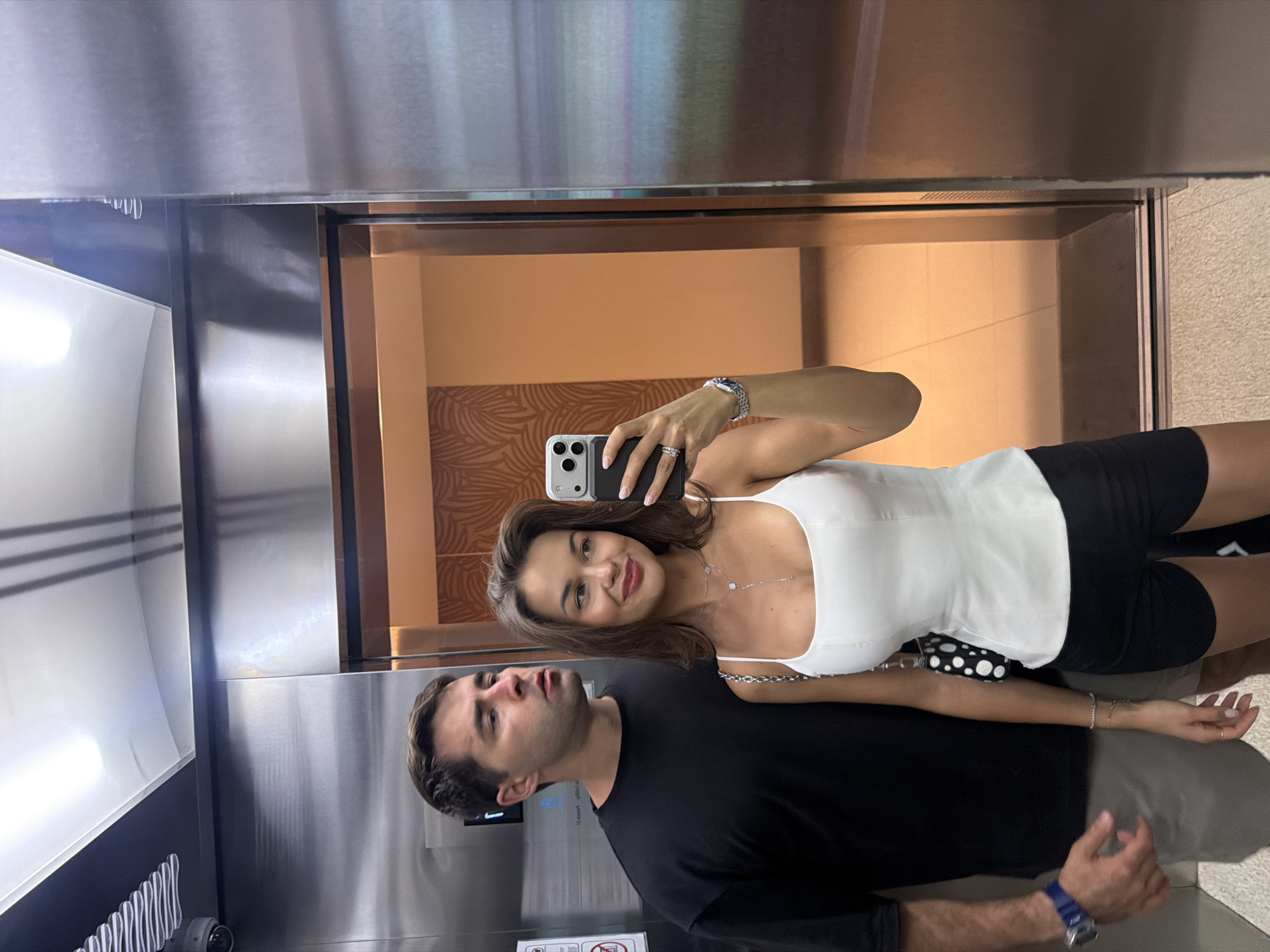 Inside an elevator in Choeng Thale, Thailand, a man and a woman pose for a selfie using a smartphone. The woman, with brown hair, is wearing a white top and black shorts, and she holds the phone. The man, wearing a black shirt and gray shorts, stands next to her. The elevator's interior is stainless steel, and a glimpse of a tiled wall can be seen through the open door behind them.